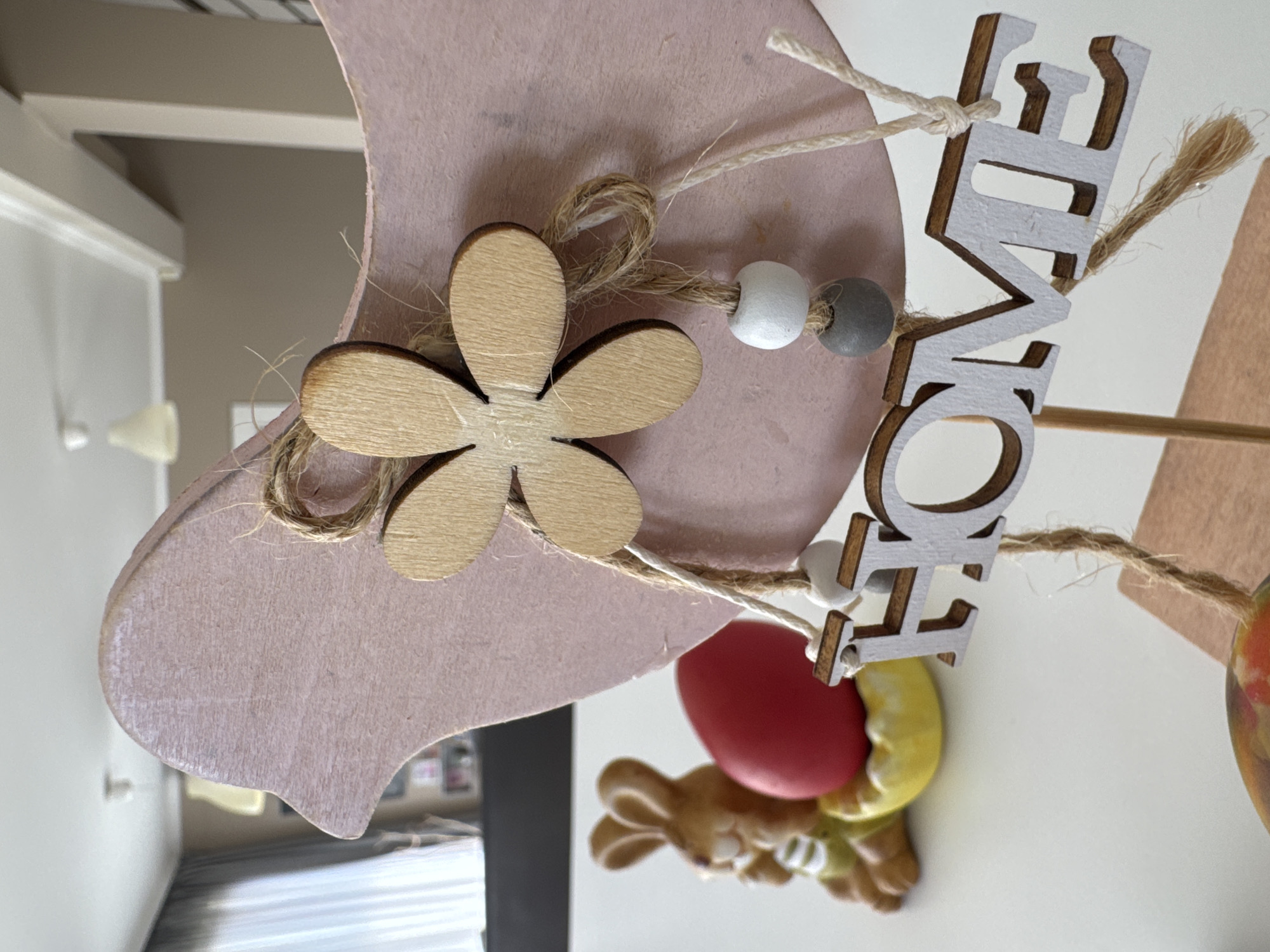 The image shows a close-up of an Easter decoration. The decoration features a pink, heart-shaped wooden piece with a wooden flower glued to it. String is tied to the decoration, with a small strand connecting to beads and the wooden letters forming the word "HOME" vertically. Below the heart, there is a ceramic bunny figurine with a red egg. The background includes a glimpse of a room with a window and a section of a white ceiling. The location in Varna, Bulgaria, cannot be determined from the visual information provided in the image.