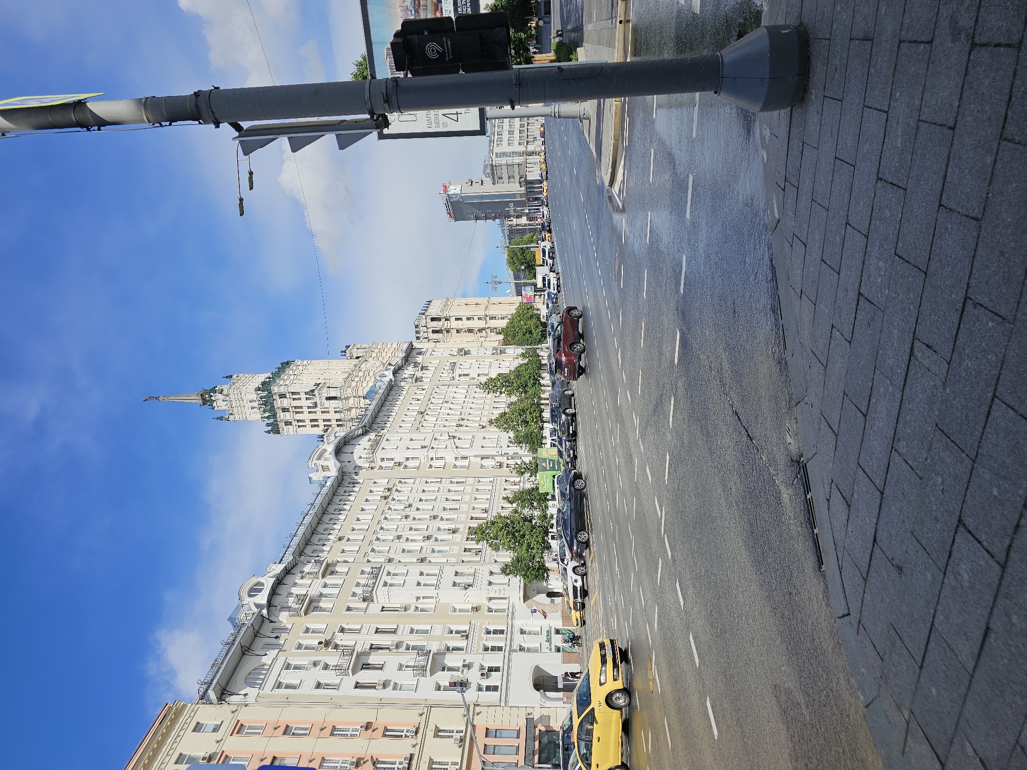 This media file depicts a street scene in Moscow, Russia, with a focus on urban architecture and vehicular traffic. The image is oriented vertically, making the road appear to run parallel to the right edge and the buildings parallel to the left edge.
The foreground features a traffic light pole and a wet asphalt road with white lane markings. Several cars are parked or driving on the road, including a prominent yellow taxi. The sidewalk to the right is paved with dark grey rectangular stones.
The background is dominated by a large, ornate building with a distinctive spire, characteristic of Stalinist architecture. Several other multi-story buildings with numerous windows and balconies line the street. Green trees are visible between some of the buildings. The sky is blue with scattered white clouds, suggesting it is daytime, likely late morning or afternoon. The wet road implies recent rain or washing.
While no people are clearly visible as main subjects, the presence of cars and taxis indicates normal city activity. The overall impression is one of a bustling urban environment. A sign with Russian text is partially visible on the traffic light pole, but it is too blurry to read. Another sign on the side of the road also contains Russian text.