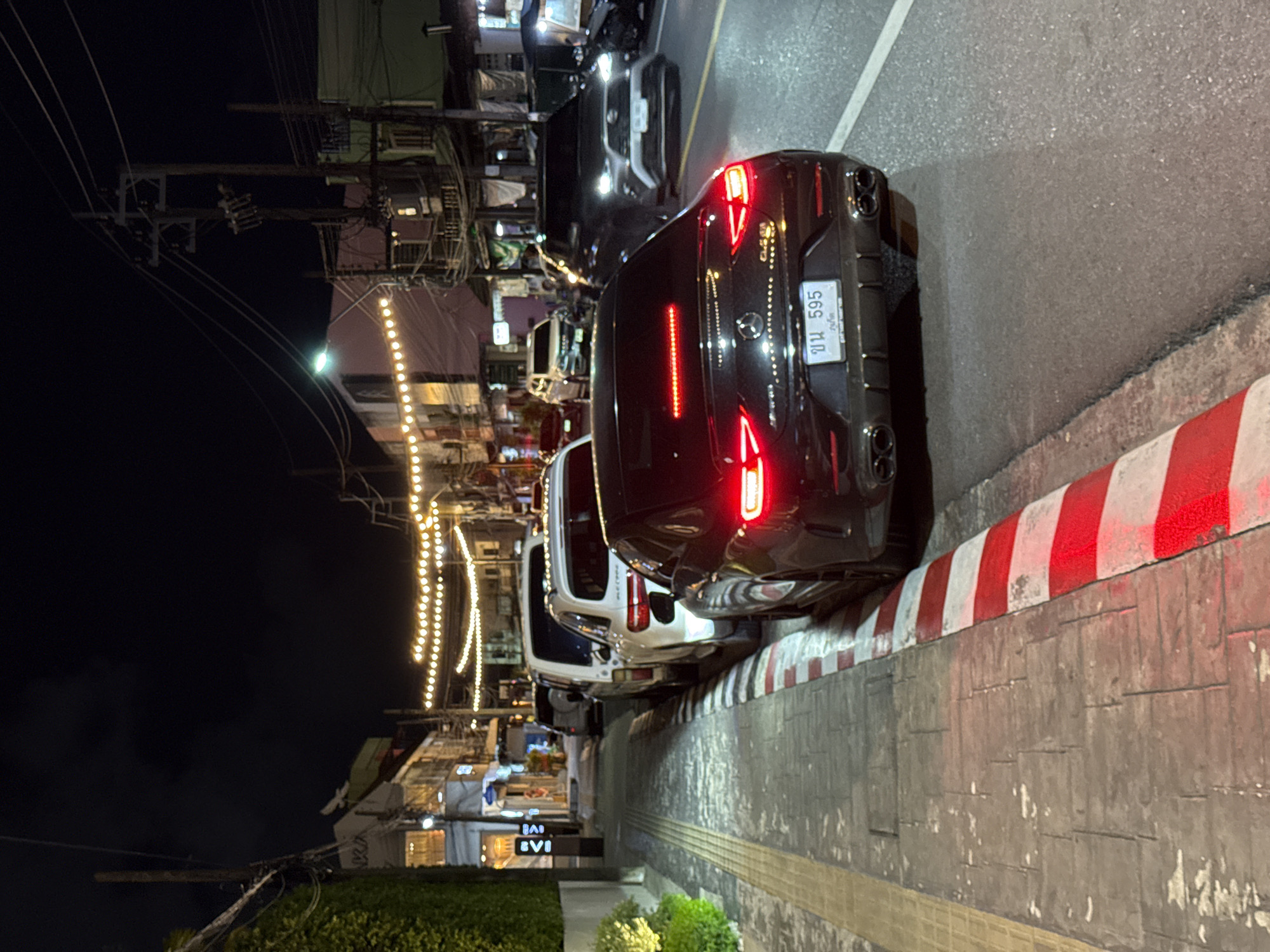 A nighttime, eye-level, full shot captures a street scene in Phuket City Municipality, Thailand, characterized by parked cars, street lights, and buildings. The foreground is dominated by the rear of a black Mercedes-Benz sedan, its brake lights illuminated. The license plate, bearing Thai characters and the number "ขบ 595", is clearly visible. To the left of the Mercedes is a red and white striped curb and a sidewalk paved with textured grey tiles. Behind the Mercedes, several white SUVs are parked along the roadside. The street itself is asphalt. Further down the street, buildings line both sides, with a string of yellow decorative lights suspended overhead, illuminating the facades. Power lines and poles are visible against the dark night sky. The overall atmosphere suggests a commercial or entertainment district at night. No people are visible in the frame.