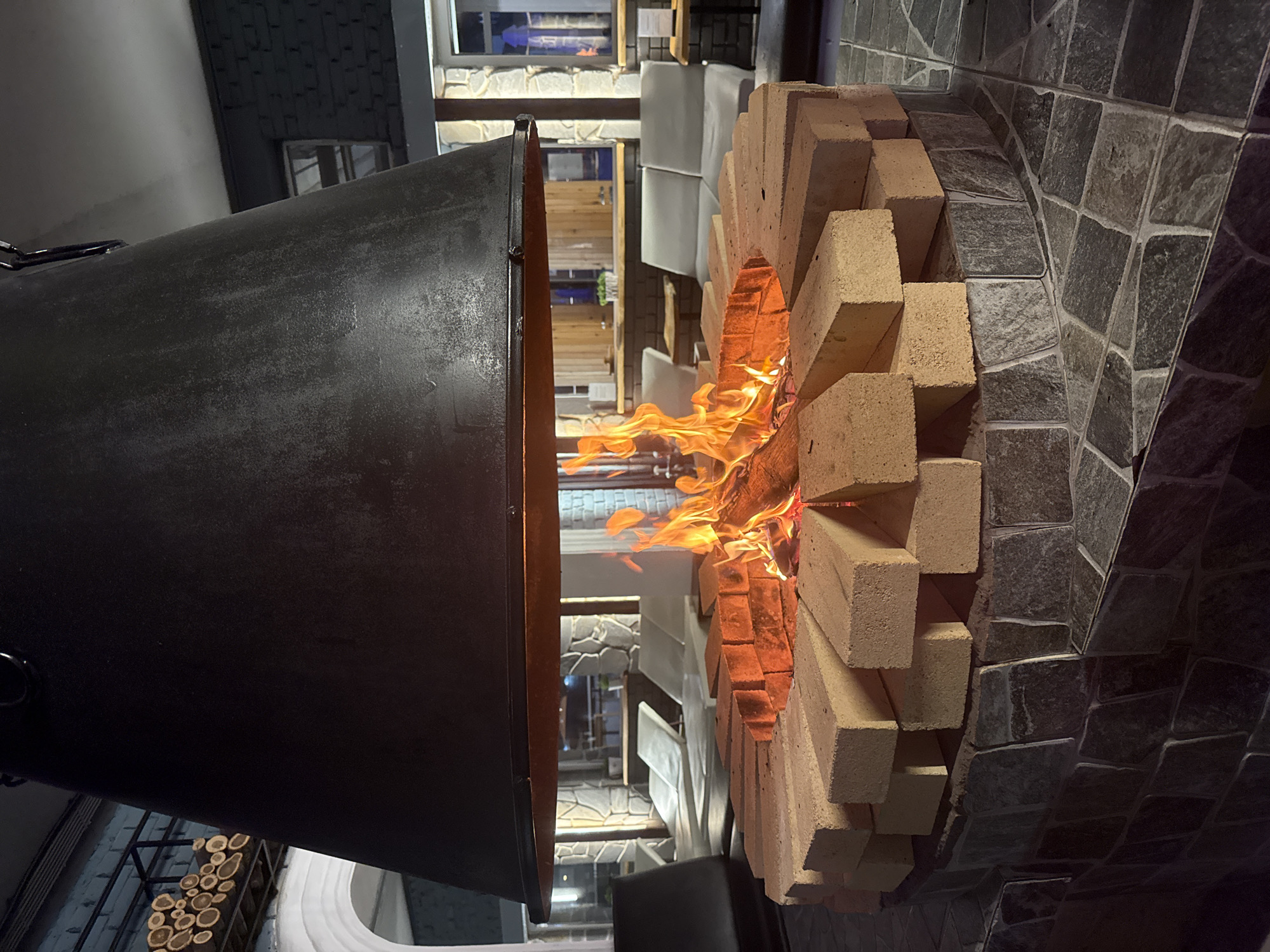 The image shows a pizza oven located in a restaurant in Rostov-on-Don, Russia. The oven is constructed of light-colored bricks and has a fire burning inside. A large metal hood hangs over the front of the oven, presumably to vent smoke and heat. The oven is set against a wall covered in dark, irregularly shaped stones. In the background, there is a view of the restaurant's interior with tables, chairs, and decorative lighting. To the left of the oven, a rack of wood sits on a shelf.