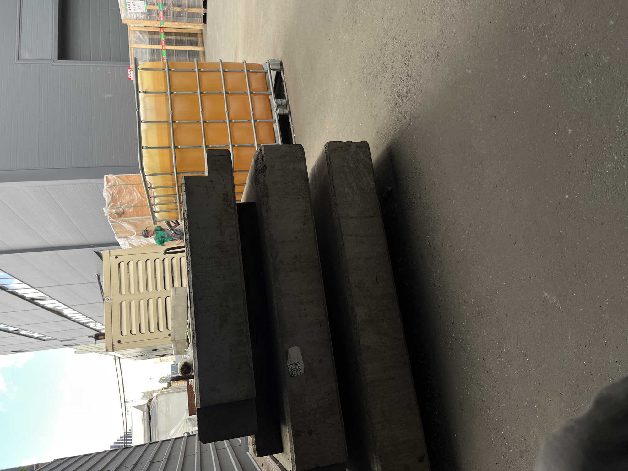 The image displays an outdoor industrial setting in Kyiv, Ukraine, with a focus on stored materials. In the foreground, several large, dark rectangular concrete slabs are stacked horizontally, casting shadows on the asphalt ground. One of the slabs has a small white sticker with a QR code and some illegible text. Behind the concrete slabs, a large, bright orange plastic IBC tote, enclosed in a metal cage, is partially visible. The tote appears to be filled with a yellow liquid. Further in the background, a beige industrial generator unit with slatted vents is situated. A person wearing a green shirt and a hard hat is visible operating a forklift behind the IBC tote. The scene suggests a storage or logistics area, possibly at a warehouse or construction site. The lighting indicates it is daytime, with clear skies visible in a cropped image. The overall impression is one of an active but orderly industrial environment.