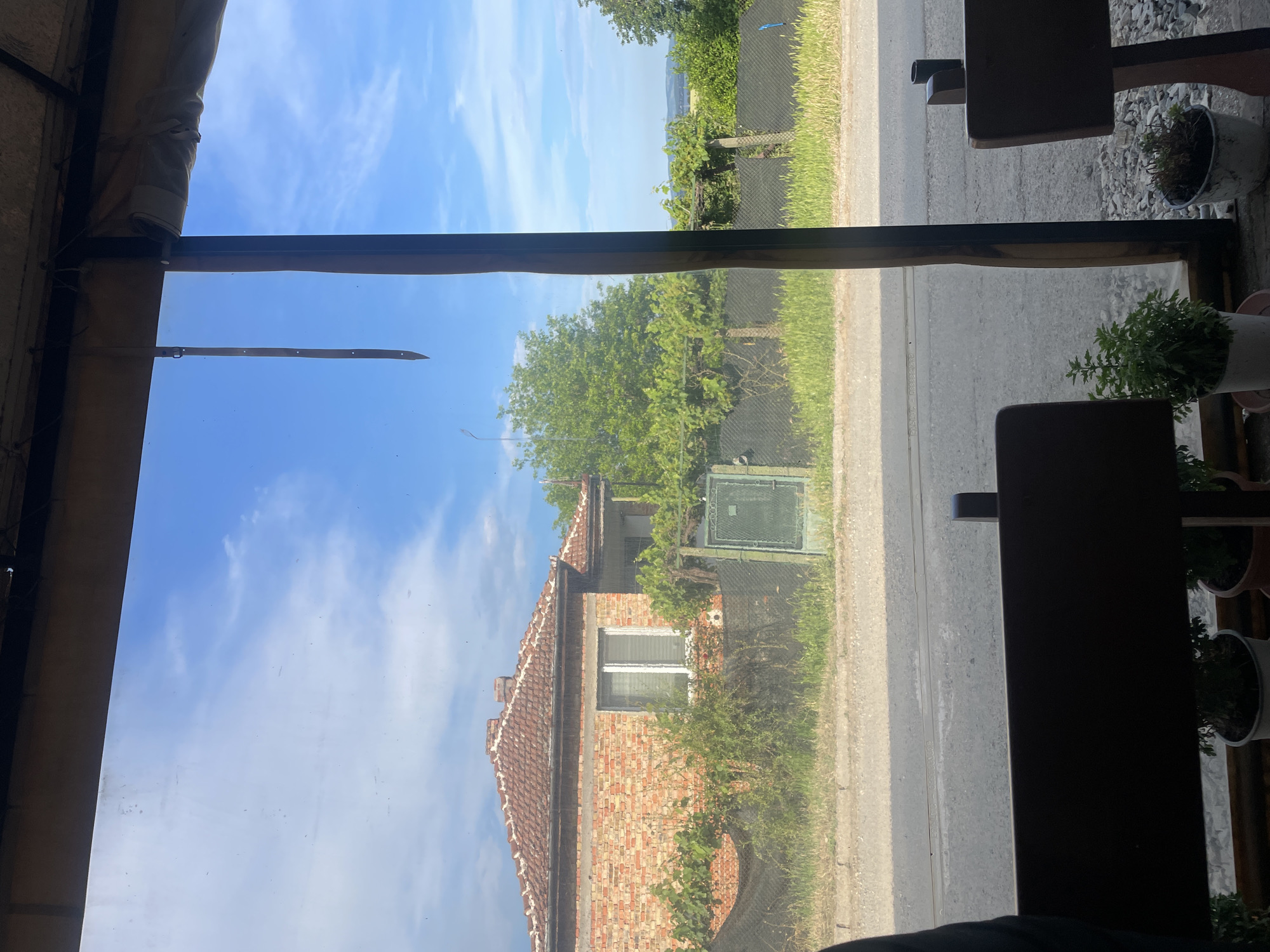 The image depicts an outdoor rural scene in Konstantinovo, Bulgaria, viewed from a sheltered interior. The top of the frame shows a beige fabric canopy structure with visible attachments; a dark brown strap with metal eyelets hangs vertically from the canopy on the upper left. A dark brown vertical post runs down the right side of the image, dividing the view. The immediate foreground, at the bottom, displays the top of a dark wooden or metal bench-like structure. Below this, three potted plants are visible: one with vibrant green foliage, and two with less distinct, possibly dried, vegetation, resting on a gravel surface.
Beyond the viewing pane, an asphalt road traverses horizontally across the middle ground. A narrow strip of green grass borders the road, followed by a dark mesh fence that extends across the scene. To the left, a single-story house with red brick walls and a red tiled hip roof is visible. The house features a prominent white-framed window with horizontal blinds. Green vines and bushes partially obscure the lower portion of the house and grow along the fence line. A small, dark green metal gate or utility box is positioned against the fence to the right of the house. Large, dense green trees populate the background behind the fence and house. The upper portion of the scene reveals a bright blue sky with scattered white clouds. Faint hills or distant terrain are visible on the horizon to the far right. No individuals or active subjects are present.