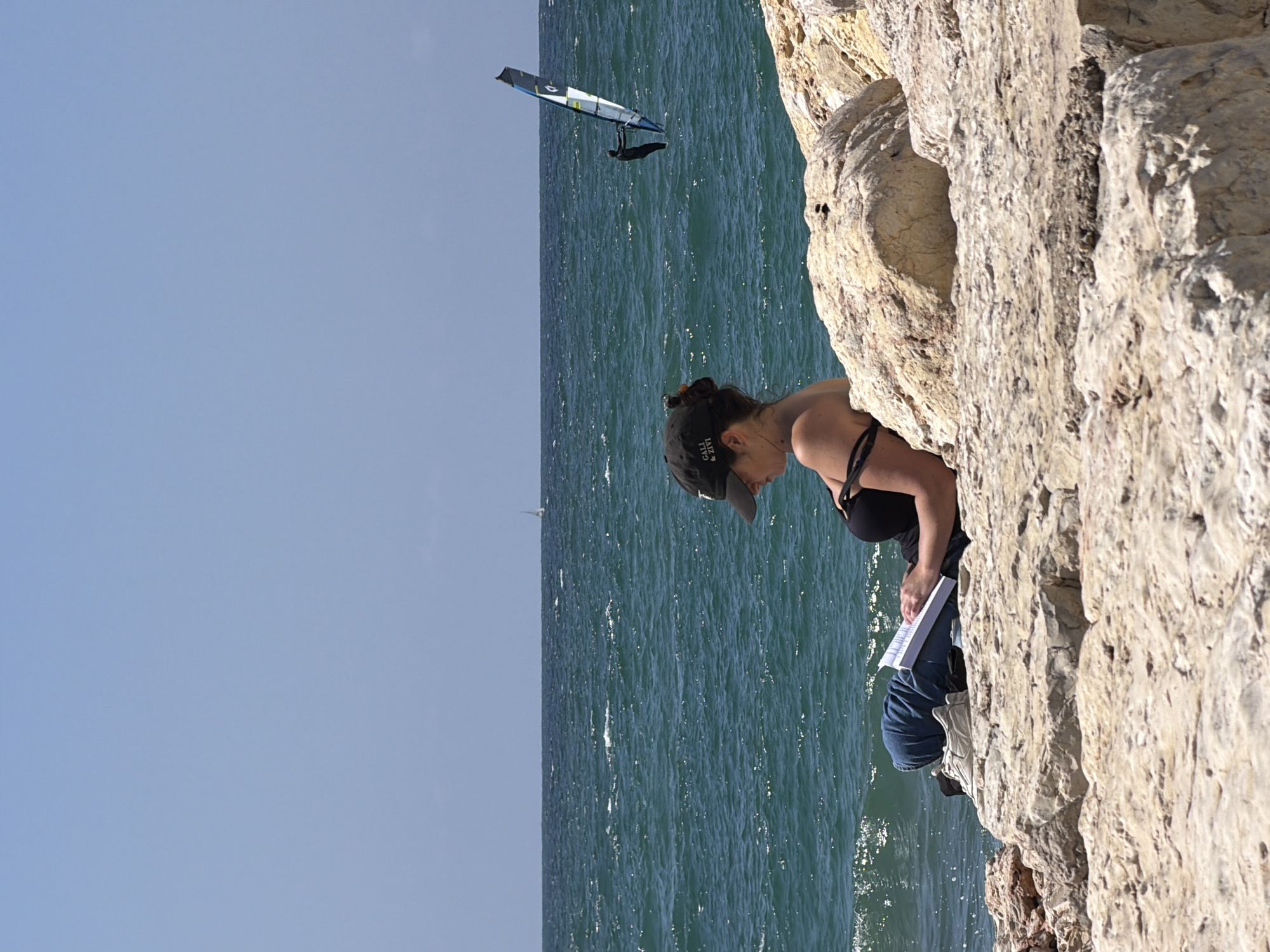 The scene is a view of the Mediterranean Sea from a cliffside location. A woman wearing a black tank top, jeans, a baseball cap, and sunglasses is sitting on the cliff, appearing to be writing or drawing in a notebook. The cliff is made of light-colored rock. In the background, the sea is visible, with a windsurfer and a small sailboat. Given the characteristics of the landscape, the image is likely to be near the beach cliffs in the Tel-Aviv area.