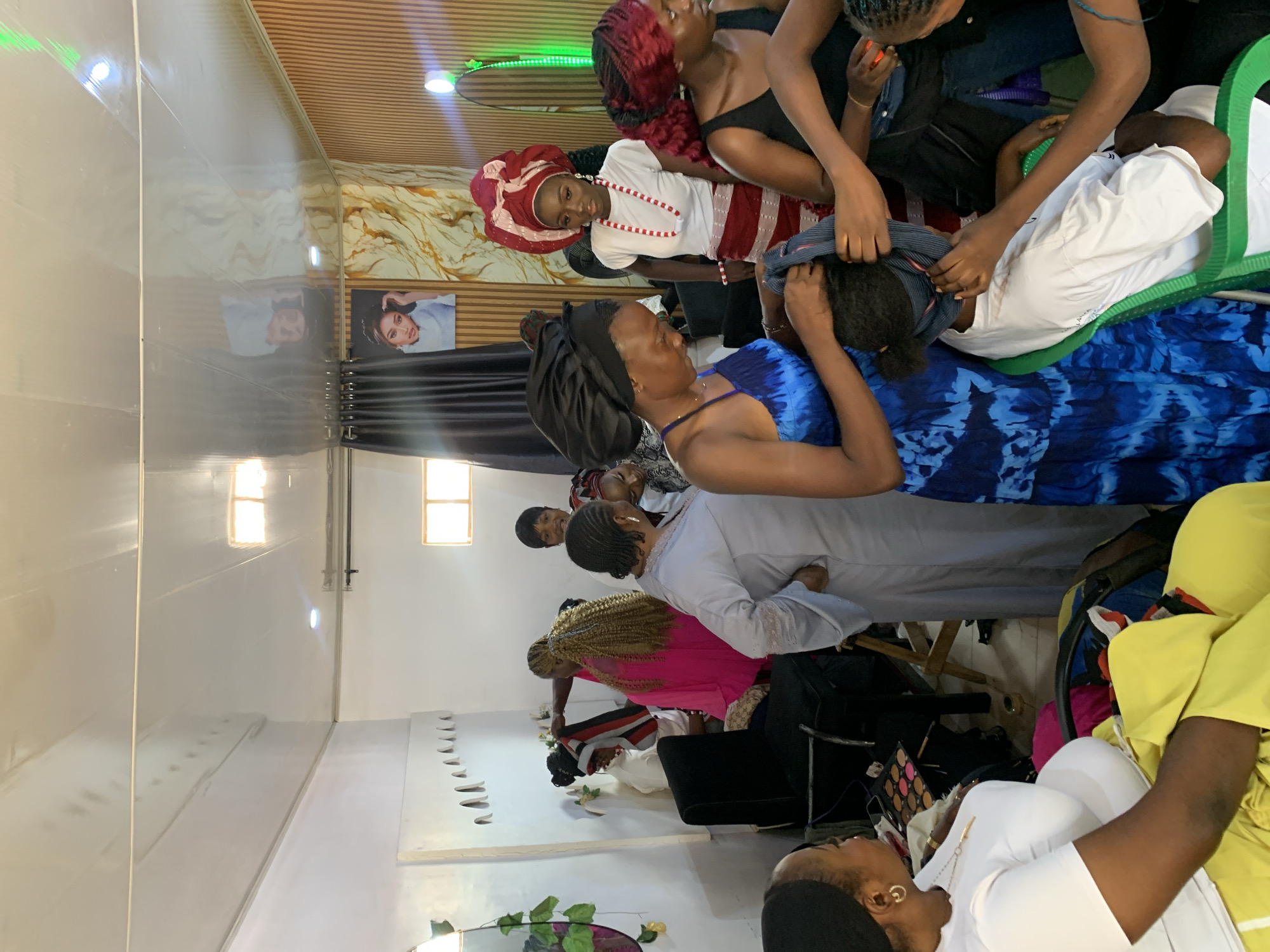The scene appears to be an indoor space, possibly a salon or dressing room, likely located in Jos, Nigeria. Several individuals are present, with one woman in a blue dress appearing to be assisting another with a headscarf or headwrap. Other women are standing around, some observing and others involved in dressing or grooming activities. A woman in traditional Igbo attire, with a white blouse and red and white skirt, is also present. Various objects such as makeup kits, bags, and clothing are scattered around the room, suggesting it is a busy and active space. The walls are painted white, and there are photographs hanging on the wall. The ceiling has a decorative wood panel design. There are bright lighting fixtures, and the overall ambiance suggests a celebratory or event preparation setting.
