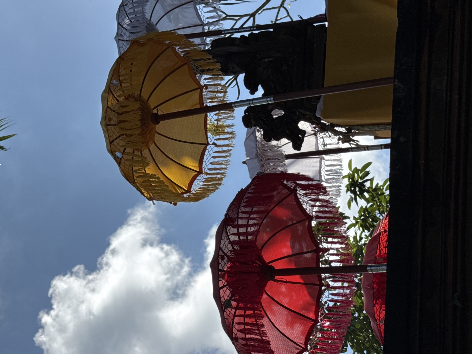 In Ubud District, Indonesia, a low-angle shot captures a festive scene beneath a bright blue sky dotted with white clouds. Several decorative umbrellas are the primary focus. In the foreground, a vibrant red umbrella is open, its intricate latticework and dangling red and white tassels visible. Behind it, another red umbrella is partially visible, also adorned with tassels. To the right and slightly further back, a large yellow umbrella stands open, its golden tassels catching the light. Behind the yellow umbrella, a white umbrella is partially visible. These umbrellas are arranged around a dark, ornate statue or shrine, which is draped in a bright yellow cloth. The overall impression is one of a cultural or religious ceremony, likely outdoors given the sky and the presence of greenery, possibly in a temple courtyard. The scene is set atop a dark, textured wall or ledge, suggesting an elevated viewpoint.