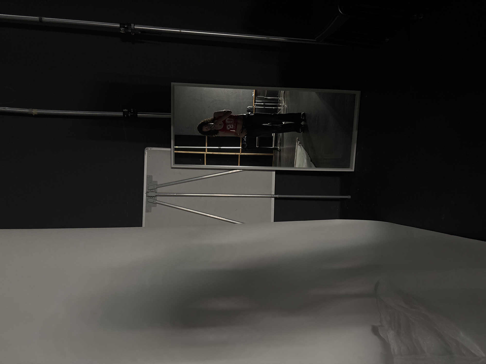 A photograph shows a person reflected in a mirror in a dark, industrial-looking room. The person is wearing a red tank top with the number "20" on it, along with dark pants. The room contains a white backdrop or screen supported by a metal stand. There are metal pipes running horizontally along the dark-painted walls. There is no visible indication of the city being Milan, Italy.