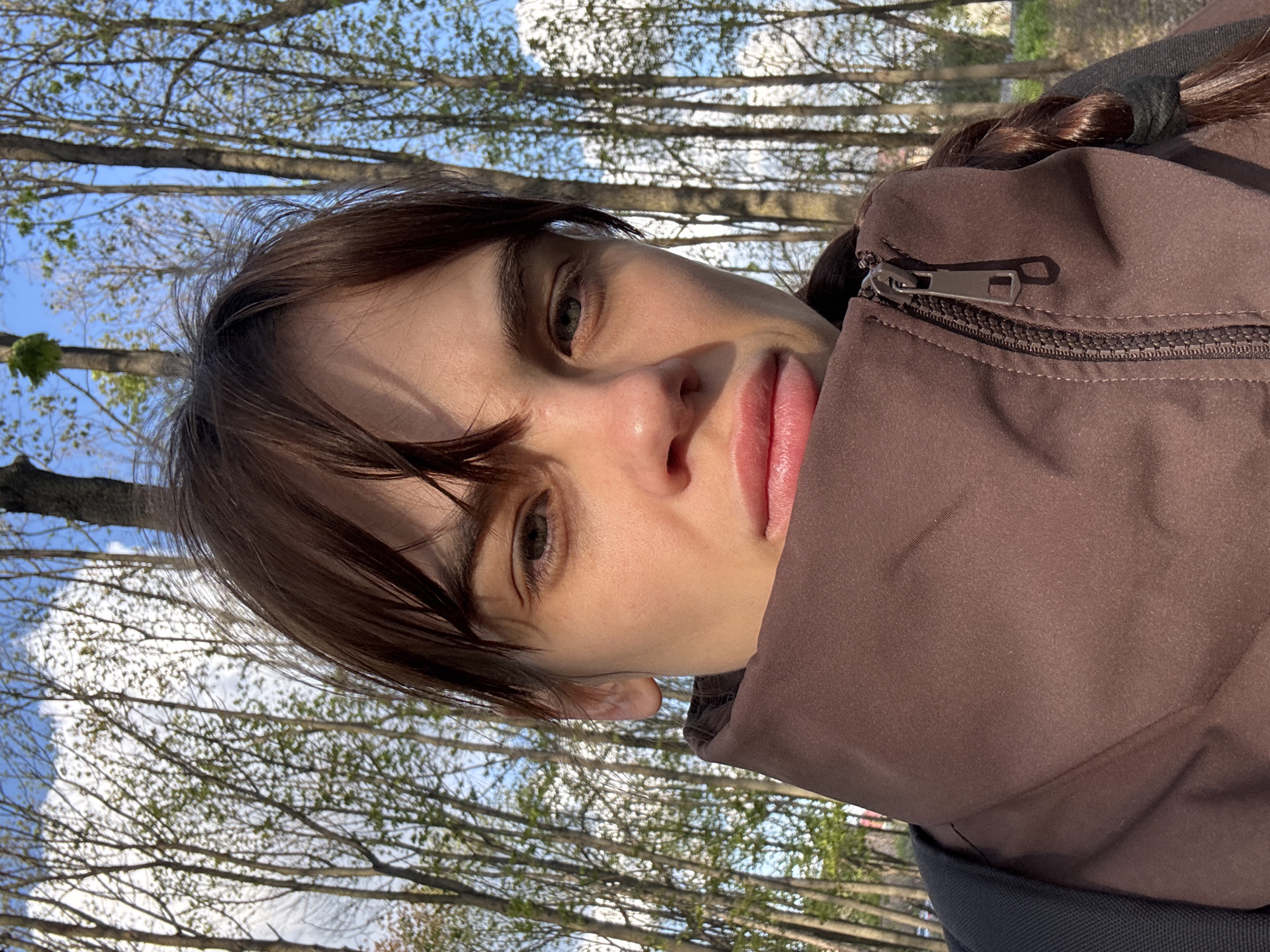 A close-up, slightly tilted, outdoor portrait of a young woman, likely in her late teens or early twenties. She has fair skin, brown hair with bangs, and is wearing a brown, puffy jacket zipped up to her neck. Her hair appears to be partially pulled back. The background consists of trees with budding leaves and a partly cloudy blue sky. The location is possibly a park or green space in a city. There is no definitive proof in this image that it is located in Rostov-on-Don, Russia.