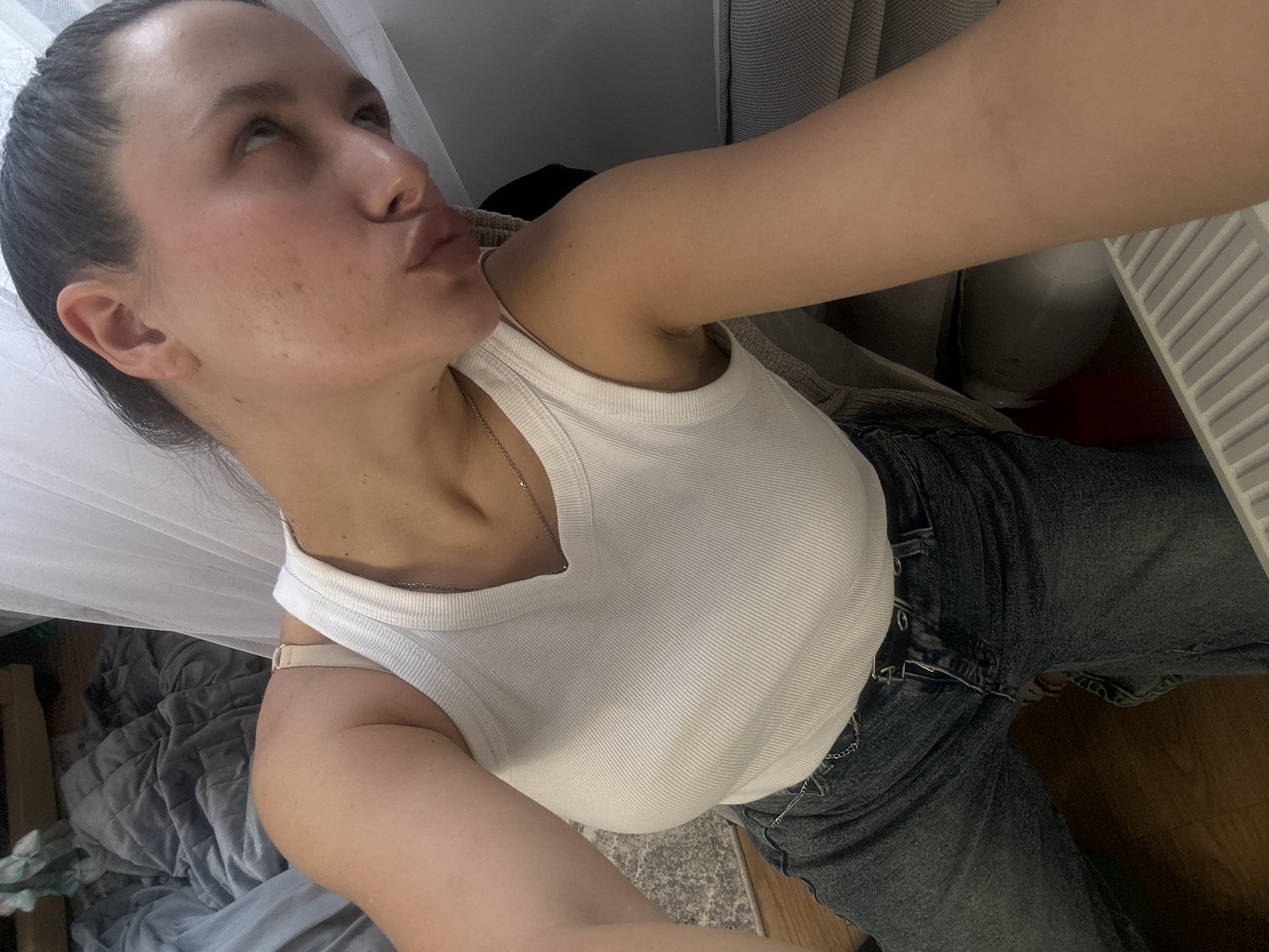 A woman is taking a selfie. She is wearing a white tank top and blue jeans. She has dark hair pulled back. The background includes a curtain, a gray blanket, and a radiator. There is no evidence to suggest this image was taken in Akulovo, Russia.