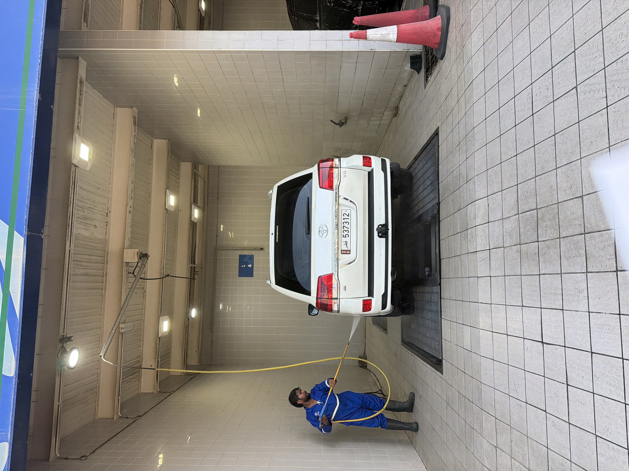 A man in a blue uniform and black boots is power washing the rear of a white Toyota Land Cruiser in a car wash bay. The car wash bay has tiled walls and a tiled floor. There are overhead lights and a yellow hose with a nozzle. In the background, there are two red and white traffic cones. The car has a license plate with the number 537312.