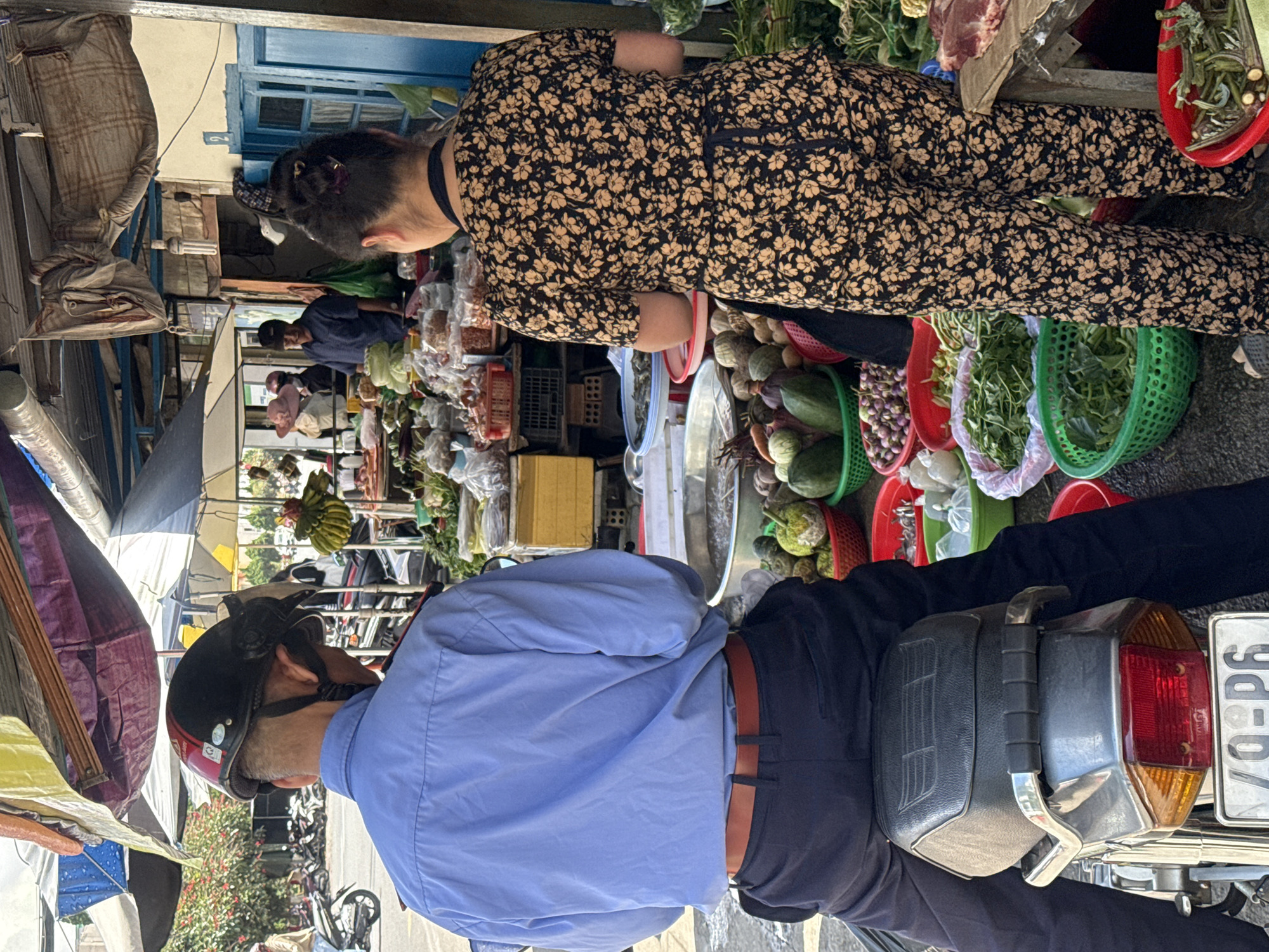 In Da Lat, Vietnam, a person wearing a blue shirt and black pants, with a black helmet on, is sitting on a motorcycle at an outdoor market. To the right of the motorcycle, another person is standing and facing away from the camera, wearing a black and floral print outfit and an apron. They are positioned next to stalls filled with produce, including leafy greens, onions, and other vegetables, as well as some meat on a table. The market appears to be under a makeshift canopy with umbrellas. In the background, other market-goers and more stalls are visible, along with parked motorcycles. The lighting suggests it is daytime.
