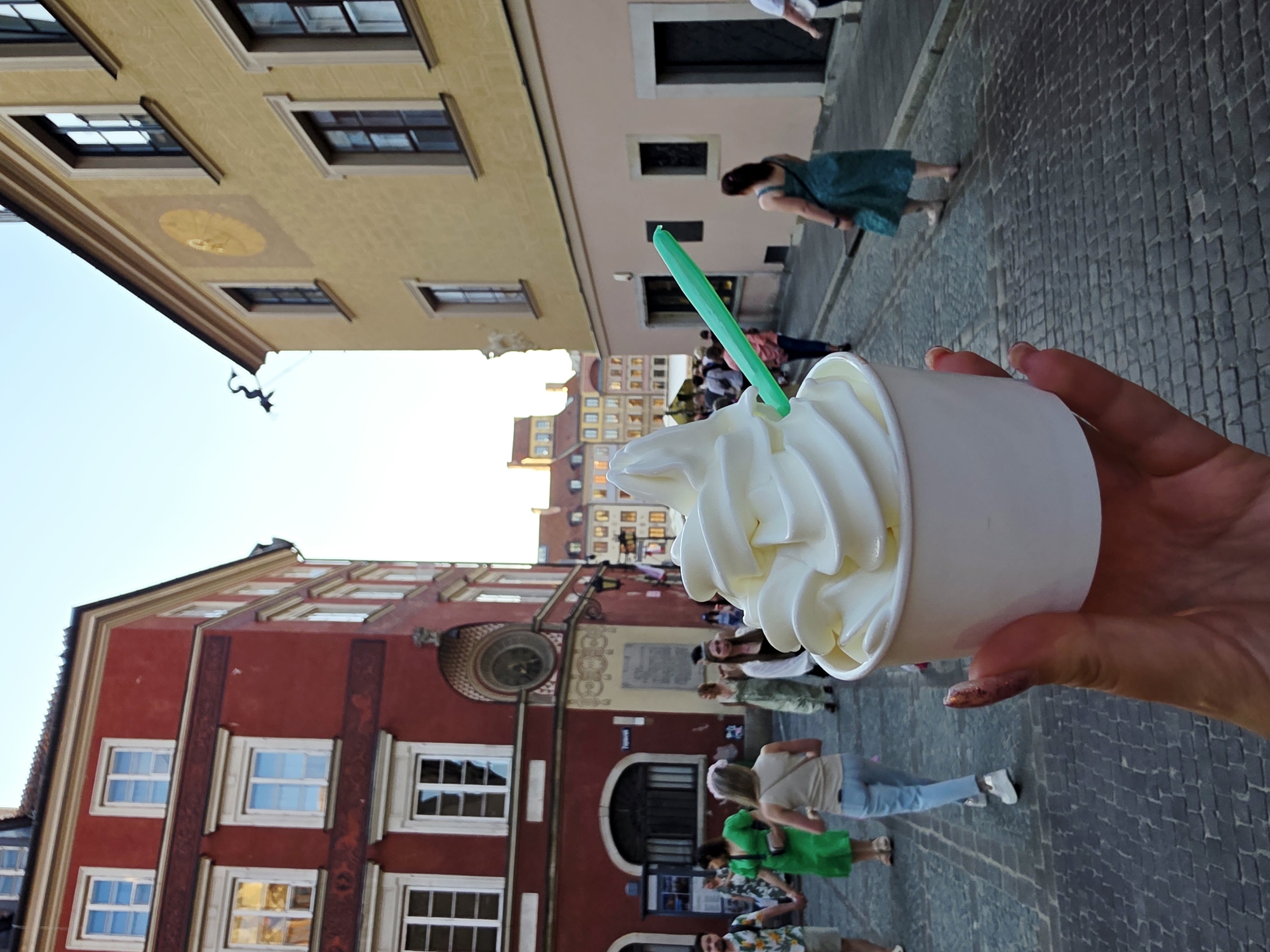 In the foreground, a person's hand holds a white paper cup filled with white soft-serve ice cream, topped with a green plastic spoon. The background depicts a street scene in Warsaw's Old Town, with cobblestone pavement. Buildings with red and yellow facades line the street, featuring windows and architectural details typical of historical European cities. Several individuals are walking in the background, some are men and women dressed in casual summer attire. The sky is clear and bright, indicating daytime.