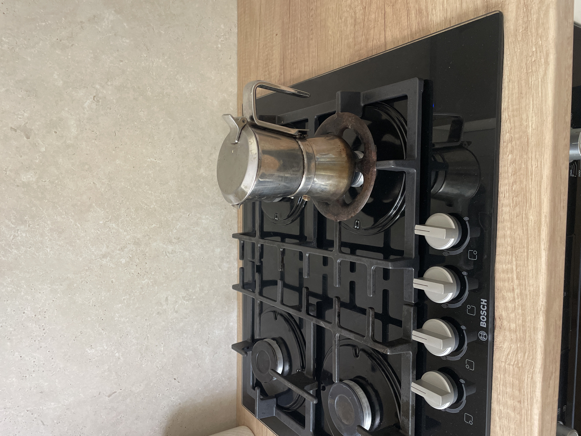 A stovetop coffee maker is situated on a black glass gas stove with a chrome finish and wooden countertop. The coffee maker is made of stainless steel and is on the right side of the stovetop. The stovetop has four gas burners and four control knobs with indicator symbols. The background is a light-colored tiled wall. The scene is set indoors, likely in a kitchen. There is no indication of the city of Cahul, Moldova in the image.