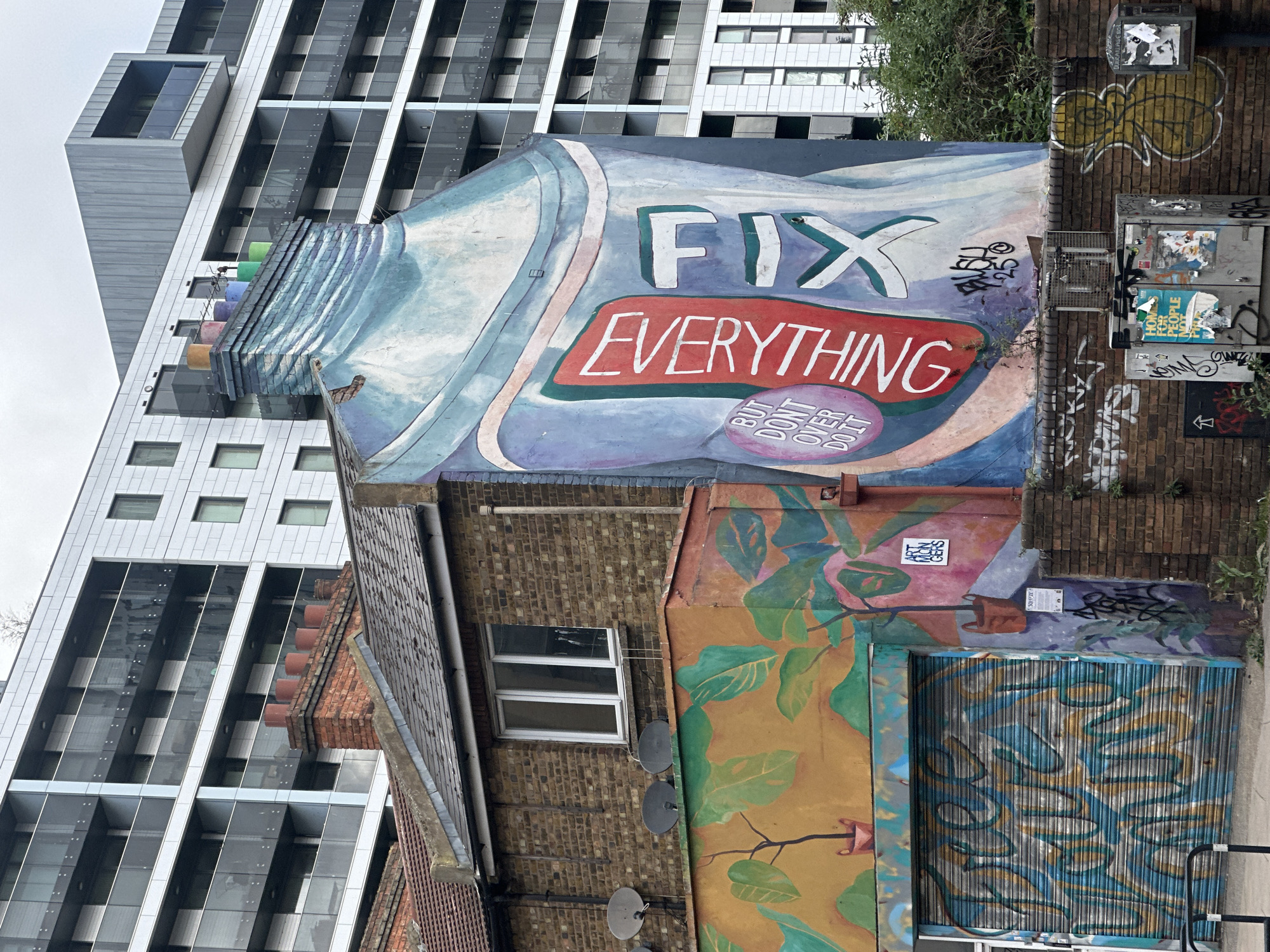 This image depicts the exterior of a building in London, United Kingdom, covered in street art. The art features the words "FIX EVERYTHING" in large white lettering over a red banner. Below this is the phrase "BUT DON'T OVER DO IT". The lower part of the building features a colorful mural with botanical themes and graffiti. A metal security shutter with abstract designs covers a section of the building's facade. Adjacent to this structure is a modern high-rise building with a glass and metal exterior. The sky is overcast. There are brick walls with additional graffiti and utility boxes with stickers and posters attached. Two satellite dishes are mounted on the brick wall. A window is visible in the brick wall.