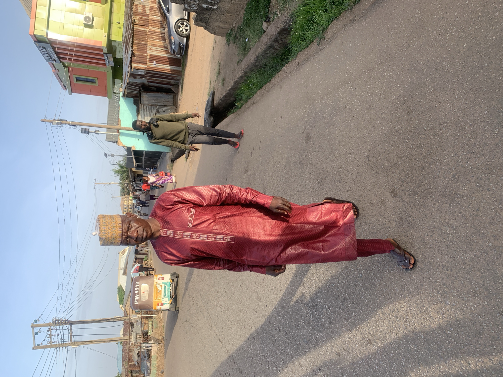 The image shows a man wearing a maroon traditional outfit and a patterned cap standing on a paved street. Another man is walking in the opposite direction. Buildings and utility poles are visible in the background, along with a vehicle with the registration number PTA 4270. The presence of this vehicle could indicate that the location is Jos, Nigeria.