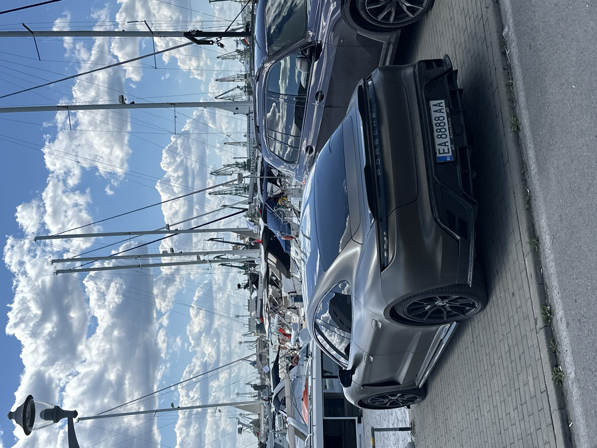 The image shows a gray Porsche car parked on a paved street in Varna, Bulgaria. The license plate reads "EA 8888 AA". A dark-colored SUV is parked behind the Porsche. In the background, numerous masts of sailboats and construction cranes are visible against a cloudy blue sky, typical of a marina or harbor scene.