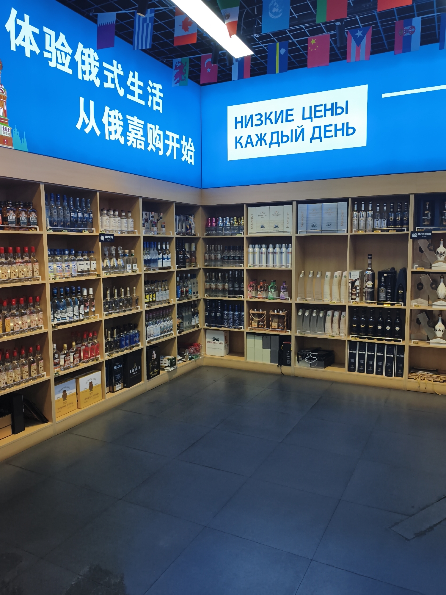 This is a medium-shot, eye-level, wide-angle photograph of the interior of a store selling alcoholic beverages. The store is brightly lit, with fluorescent lights overhead and illuminated signage. The signage at the top of the image displays white Chinese characters on a bright blue background, and also features a white rectangle with blue Cyrillic text. Several small national flags are strung across the top of the display.
The store is organized with a series of wooden shelving units filled with bottles of liquor. The shelves are arranged in a grid pattern, with numerous individual compartments. The bottles vary in size, shape, and color, with many clear bottles containing clear liquids, and others containing colored liquids. Some bottles are stacked horizontally, while others are displayed vertically. There are also some boxes and decorative items on the shelves.
The floor of the store is tiled with large, dark gray square tiles. The overall impression is that of a well-stocked liquor store with a modern and clean aesthetic. The store's inventory suggests it might be catering to an international clientele, given the various flags and the Cyrillic text on the signage.
This image was taken in Xi'an, China, specifically at the "Siberian Liquor Store" located in the "Xian International Trade City" (西安国际贸易城).
