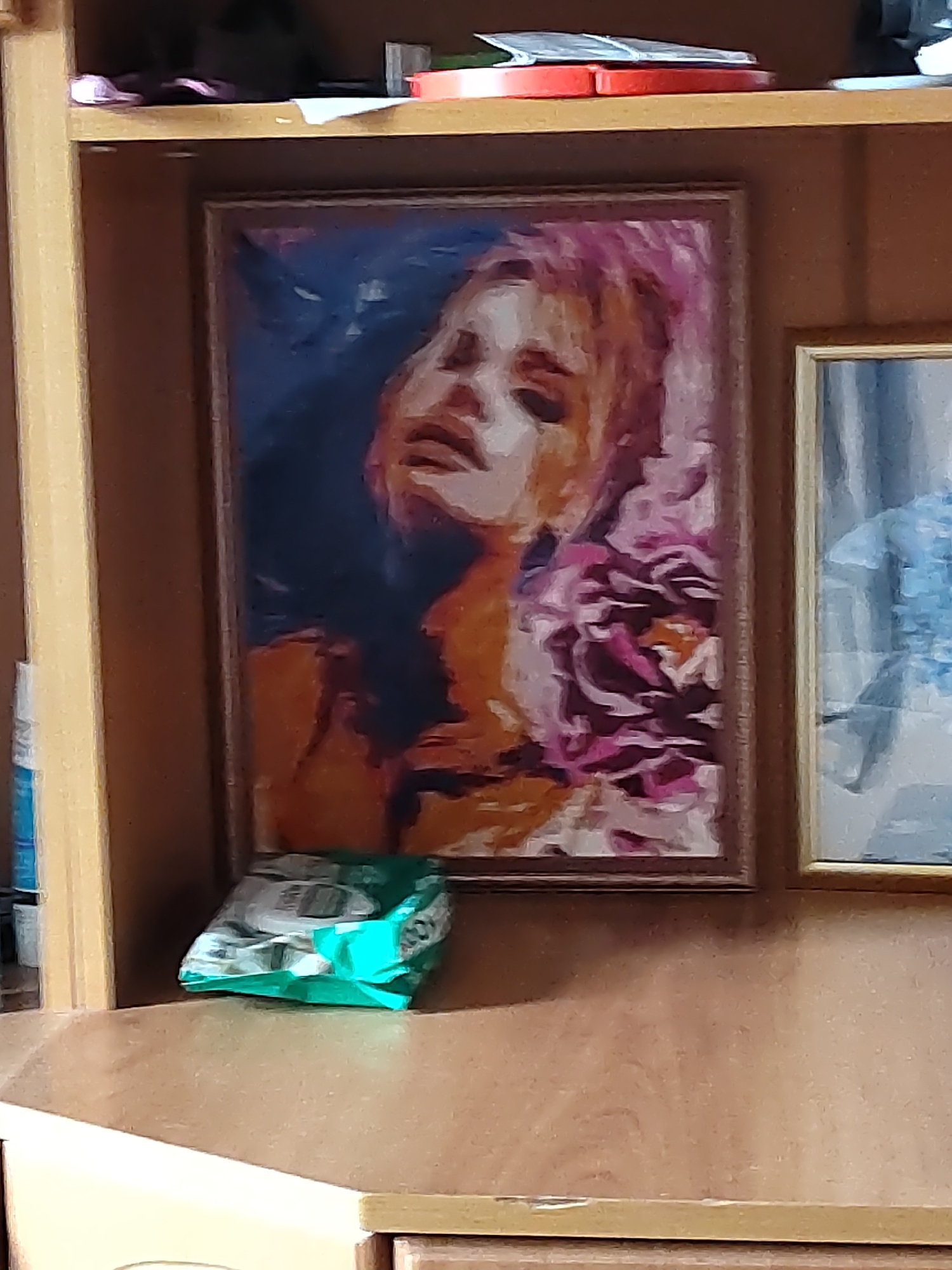 The image shows a close-up of a wooden shelf unit in what appears to be a domestic setting. On the top shelf, there are various items including what looks like a red object, possibly a book or a box, and some white papers. Below this, a framed painting of a woman's face with pink hair is displayed prominently. To the right of this painting, another framed painting, largely out of focus and depicting blue tones, is visible. In front of the larger painting, on the shelf below, sits a crumpled green plastic bag containing some items. The environment suggests a home or storage area, possibly in Бубеньево, Russia, given the context. The lighting is somewhat dim, creating shadows and highlighting the texture of the wood and the painted surfaces.