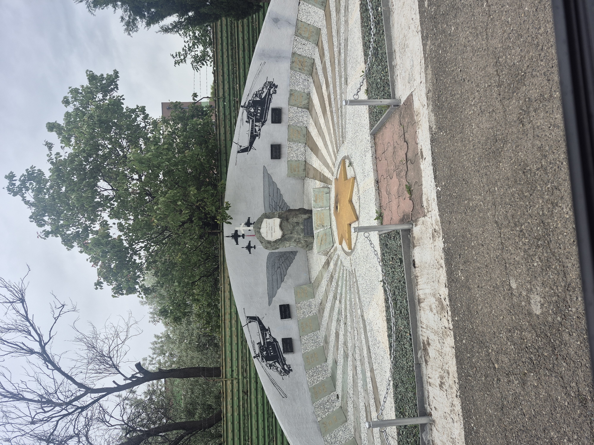 The image shows a war memorial located in Tbilisi, Georgia. The memorial is semicircular and features a central star-shaped symbol. The memorial has drawings of helicopters and fighter planes on the left side and is bordered by a metal railing. Trees are visible in the background, and a paved surface is to the right of the memorial. The sky is overcast.