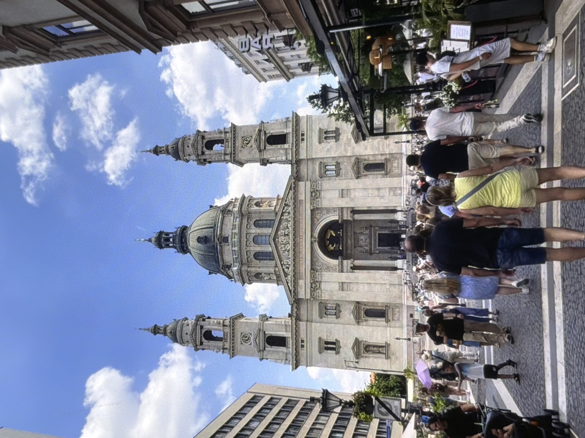 The image captures a street scene dominated by a large, ornate building, likely a church or cathedral, with prominent towers and a dome. The architecture appears classical, with intricate stonework and numerous windows. A portion of a modern building with a grid-like facade is visible in the foreground.
A crowd of people are gathered on the street and sidewalk in front of the grand building, suggesting a tourist or public gathering. They are dressed in casual, warm-weather clothing, indicating it is daytime and likely a pleasant day. Some individuals are walking, while others appear to be observing the architecture or conversing. A few people are looking at their phones. One person is holding a purple umbrella.
The sky is blue with scattered white clouds. The lighting suggests it is daytime, with the sun casting some shadows. The scene has a lively atmosphere, with many people present. There is a sign on the modern building in the upper left corner with the letters "BAR" clearly visible. The overall setting is urban, with elements of historical architecture juxtaposed with more contemporary structures.
