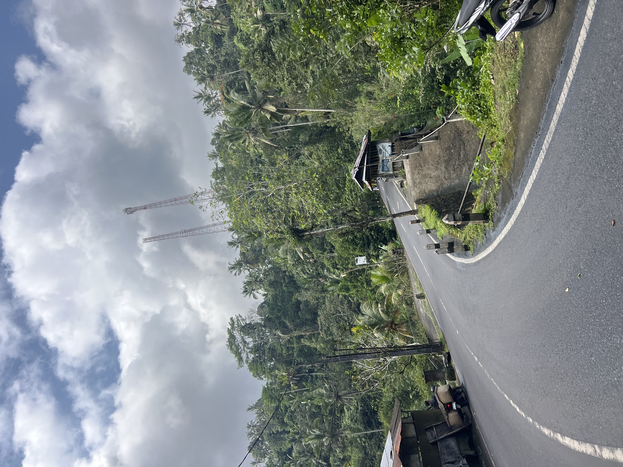 The image depicts a winding asphalt road through a lush, tropical hillside, likely in Tembuku, Indonesia. The road curves from the foreground to the left, bordered by a steep, overgrown embankment on the right. Several utility poles with wires run along the left side of the road. A small, rustic wooden structure with a corrugated metal roof is situated on the right side of the road, elevated slightly from the street level. Two tall, red and white communication towers are visible in the distance, rising above the dense green foliage of palm trees and other tropical vegetation that covers the hillside. In the foreground on the left, a motorcycle is parked next to a simple, open-sided structure made of corrugated metal and wood. A person is seated on the motorcycle, which is loaded with woven baskets. The sky is partly cloudy with patches of blue sky showing through the dense, grey clouds.