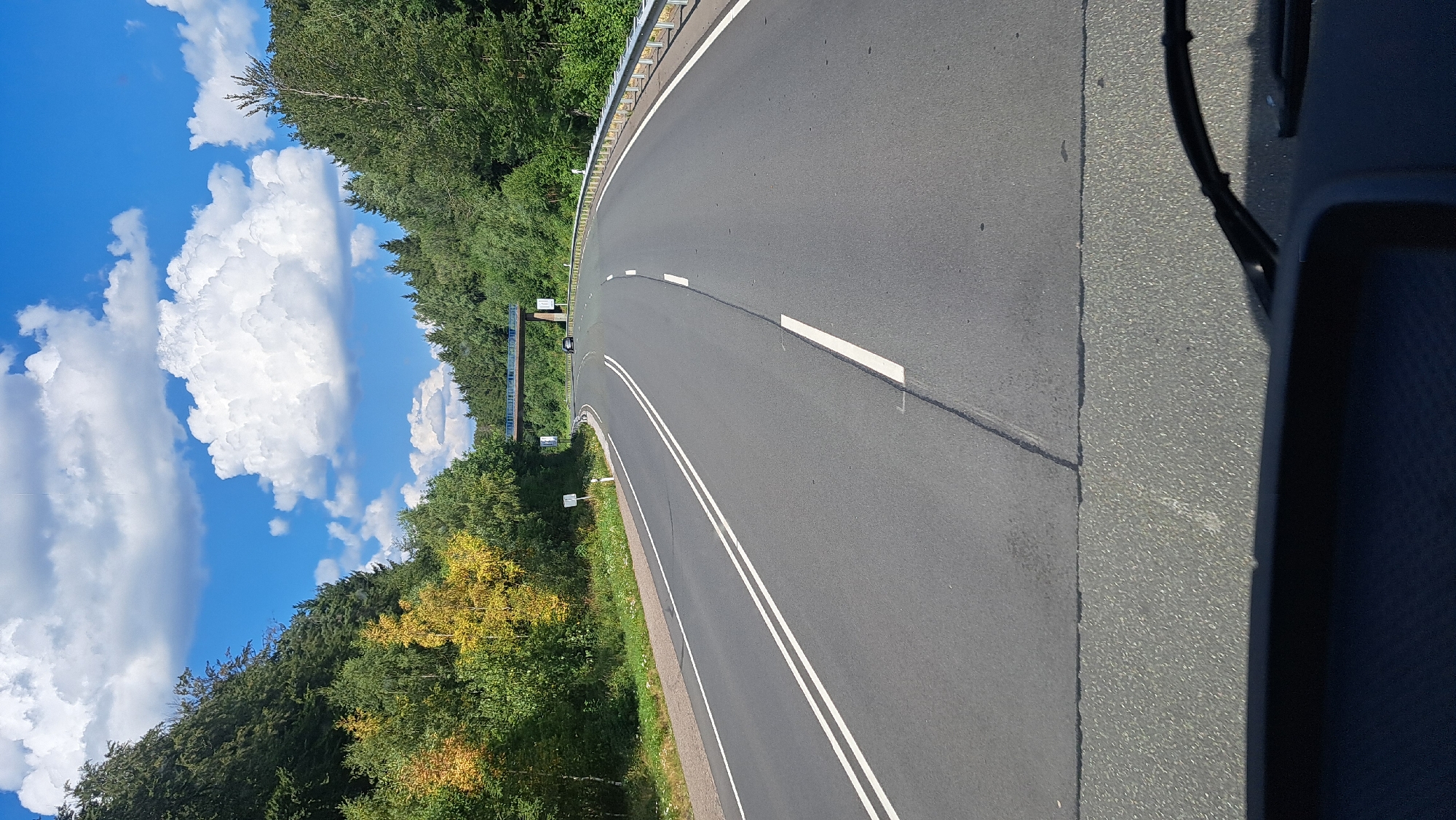 The image shows a view from inside a vehicle driving on a paved road in a forested area. The road curves to the right and has two white lines in the center, indicating a two-lane road. There is a guardrail on the right side of the road. In the distance, there is a bridge crossing over the road, and a few cars are visible on the bridge. The sky is blue with white, fluffy clouds. The trees on either side of the road are green, with some yellow foliage on the left side, suggesting it may be early autumn. The foreground shows the dashboard and windshield wipers of the vehicle. This scene is consistent with a road in the vicinity of Burbach, Germany, a town known for its scenic natural surroundings and forested landscapes.