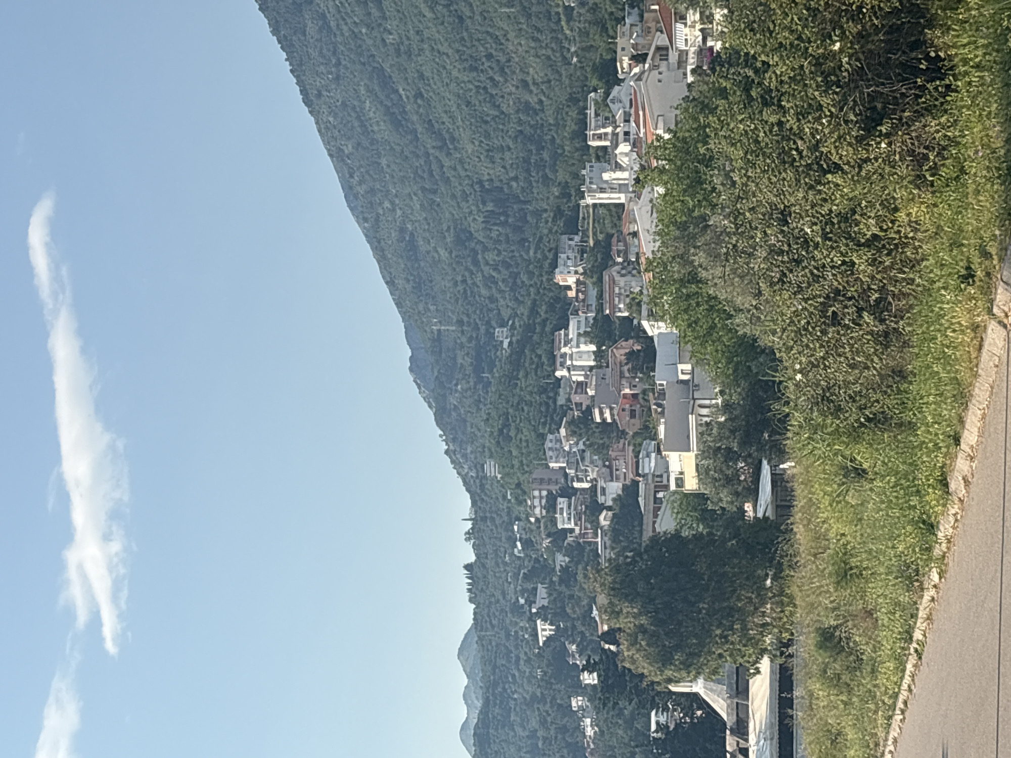 A daytime shot shows a mountainside community in Bar, Montenegro. The left side of the frame reveals a steep, tree-covered mountain. To the right, a dense collection of houses and buildings dot the slope, appearing to cascade down the mountain. Lush green vegetation borders a paved road on the right-hand side. The sky is a clear blue with a wisp of a cloud on the left. The image seems to have been taken from ground level, capturing the scene at a slight angle.