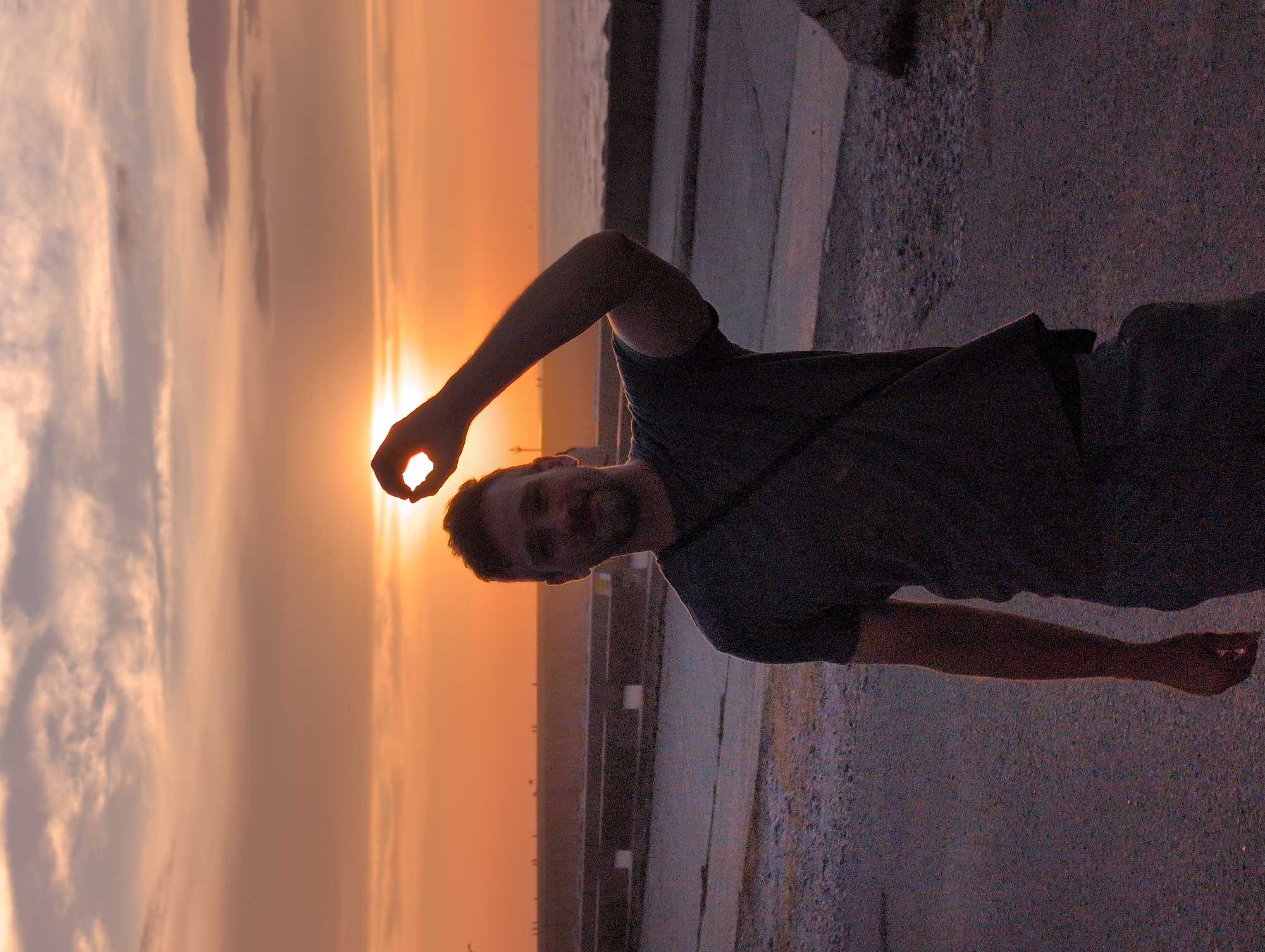 A man stands outdoors at dusk, silhouetted against a warm sunset. He is holding his left hand up, with his thumb and index finger forming a circle through which the sun is visible. The man is wearing a dark t-shirt and dark pants. He has short, dark hair and a beard. The background shows the sea and a concrete promenade or pier. The sky is filled with wispy clouds, tinged with orange and pink from the setting sun. The ground in front of him is an asphalt surface. The scene suggests a coastal location, likely in the town of Le Tréport, France, known for its dramatic cliffs and harbor.