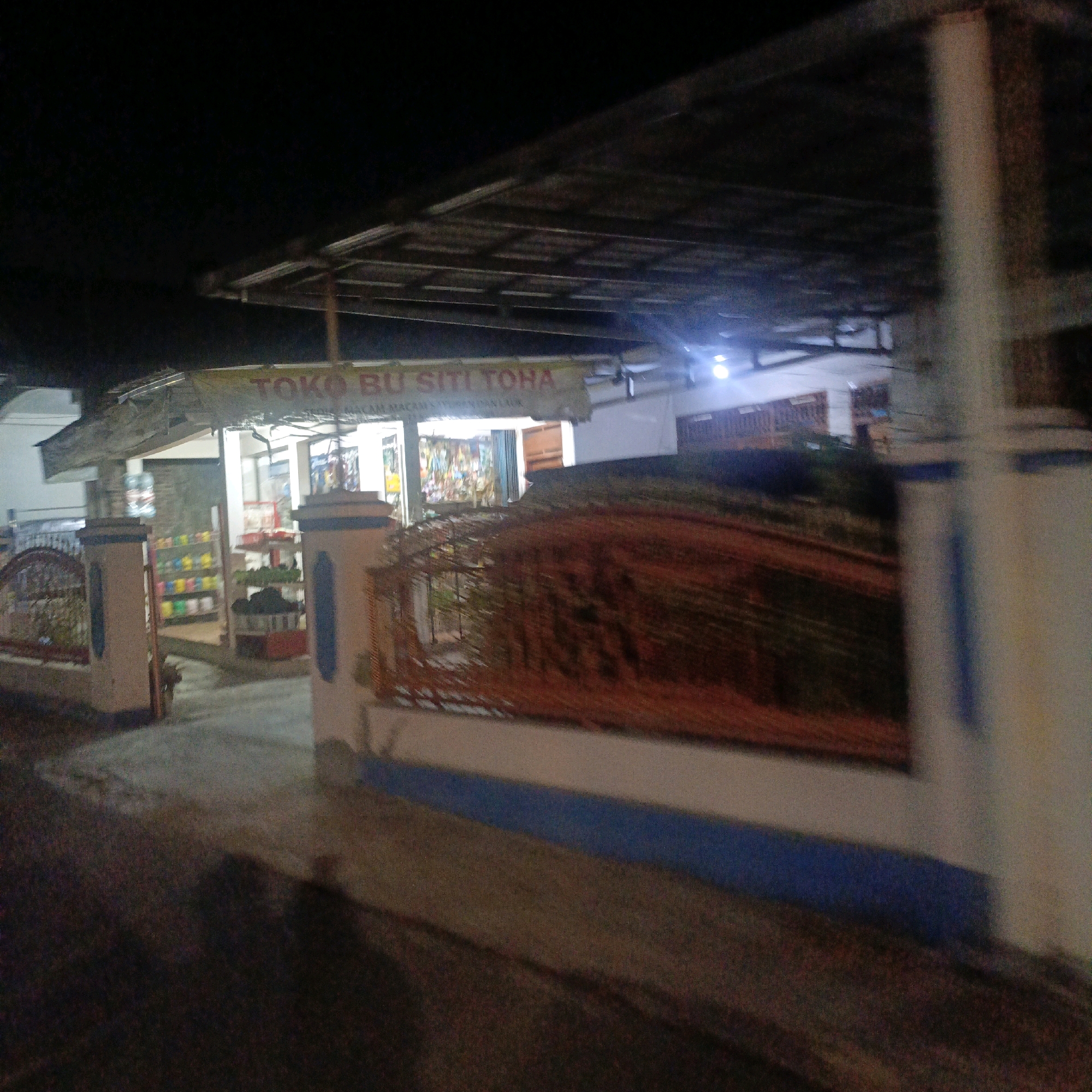 The image shows a nighttime view of a storefront in Salatiga, Indonesia. A banner above the entrance reads "TOKO BU SITI TOHA" in yellow letters, with smaller text below it that appears to be a description of the goods sold. The store appears to be a small, local shop selling various items, as evidenced by the shelves stocked with colorful products visible through the open doorway and windows. The storefront has an overhang supported by poles, and a decorative fence with a red, woven material runs along the front of the property. The lighting is dim, suggesting it is late at night, and there are no people clearly visible in the image. The overall impression is of a typical Indonesian neighborhood store.