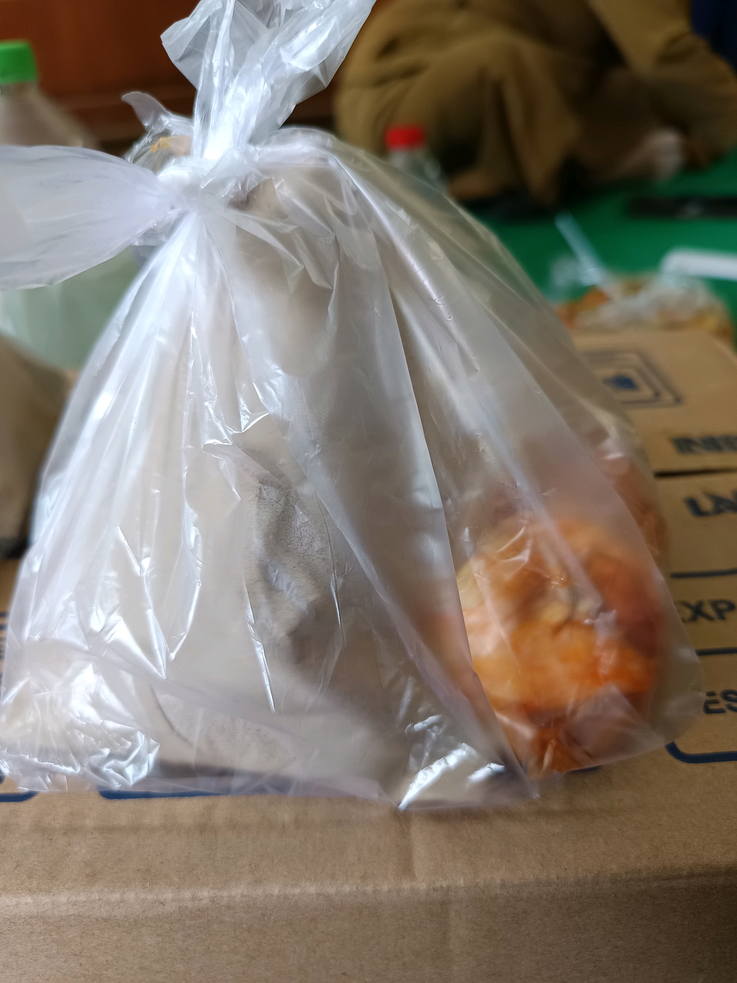 The image shows a plastic bag containing food, likely takeout, placed on a cardboard box. The bag is clear plastic and tied at the top. Inside the bag, a light-colored, powdery substance is visible on the left, and an orange-colored food item, possibly fried chicken or pastries, is seen on the right. The background is blurred, but suggests an indoor setting with some other objects and a green surface. The scene appears to be a close-up shot focusing on the food bag. The location is not identifiable from the image itself.