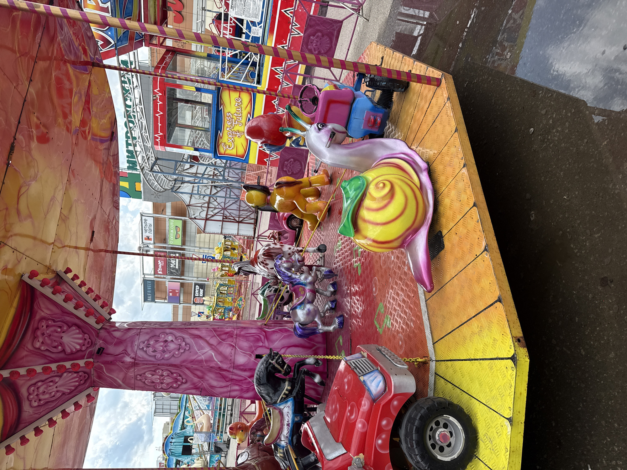 The image shows a children's carousel ride in Dmitrov, Russia. The carousel features various animal figures and vehicles for children to sit on, including a snail, a horse, and a red car. In the background, buildings are visible, and signs for businesses, including a burger restaurant ("Бургер Кинг"), a pet store ("четыре лапы"), and a sign that translates to "Express of Future." The ground near the carousel appears wet and dirty.