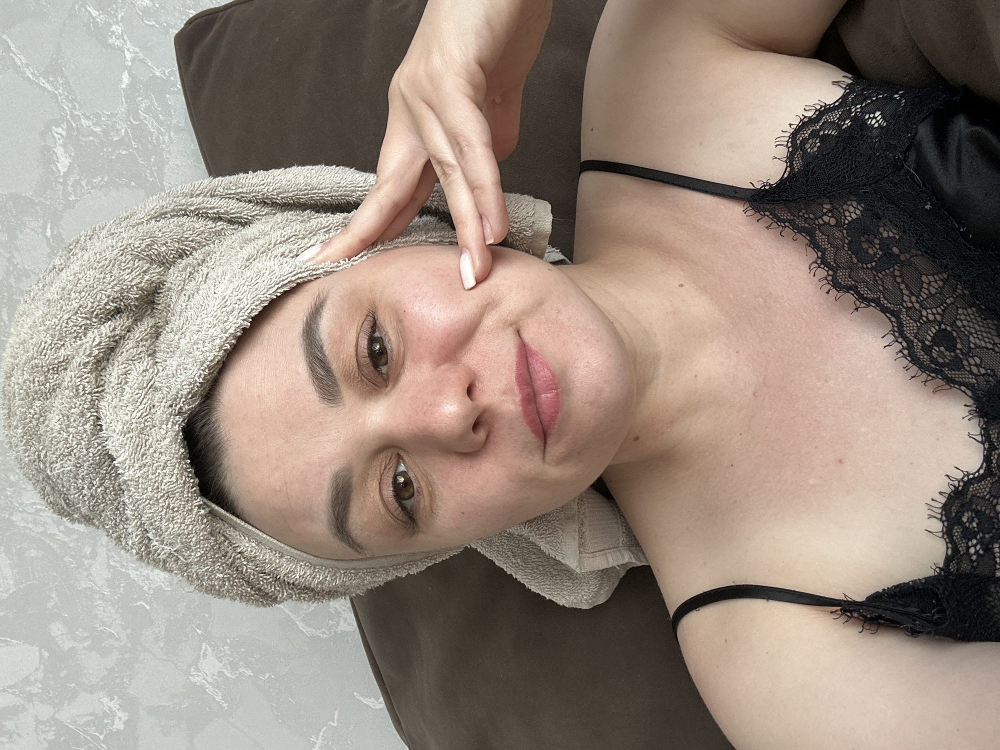 A selfie, possibly taken in Chaikovsky, Russia, shows a woman lying on a brown surface, likely a pillow or cushion. She wears a towel wrapped around her head and a black lace-trimmed top with thin straps. Her left hand is near her forehead, with fingers gently touching her skin. Her face is angled towards the camera, showing her eyes, nose, lips, and eyebrows. The background includes a wall with a marbled pattern.
