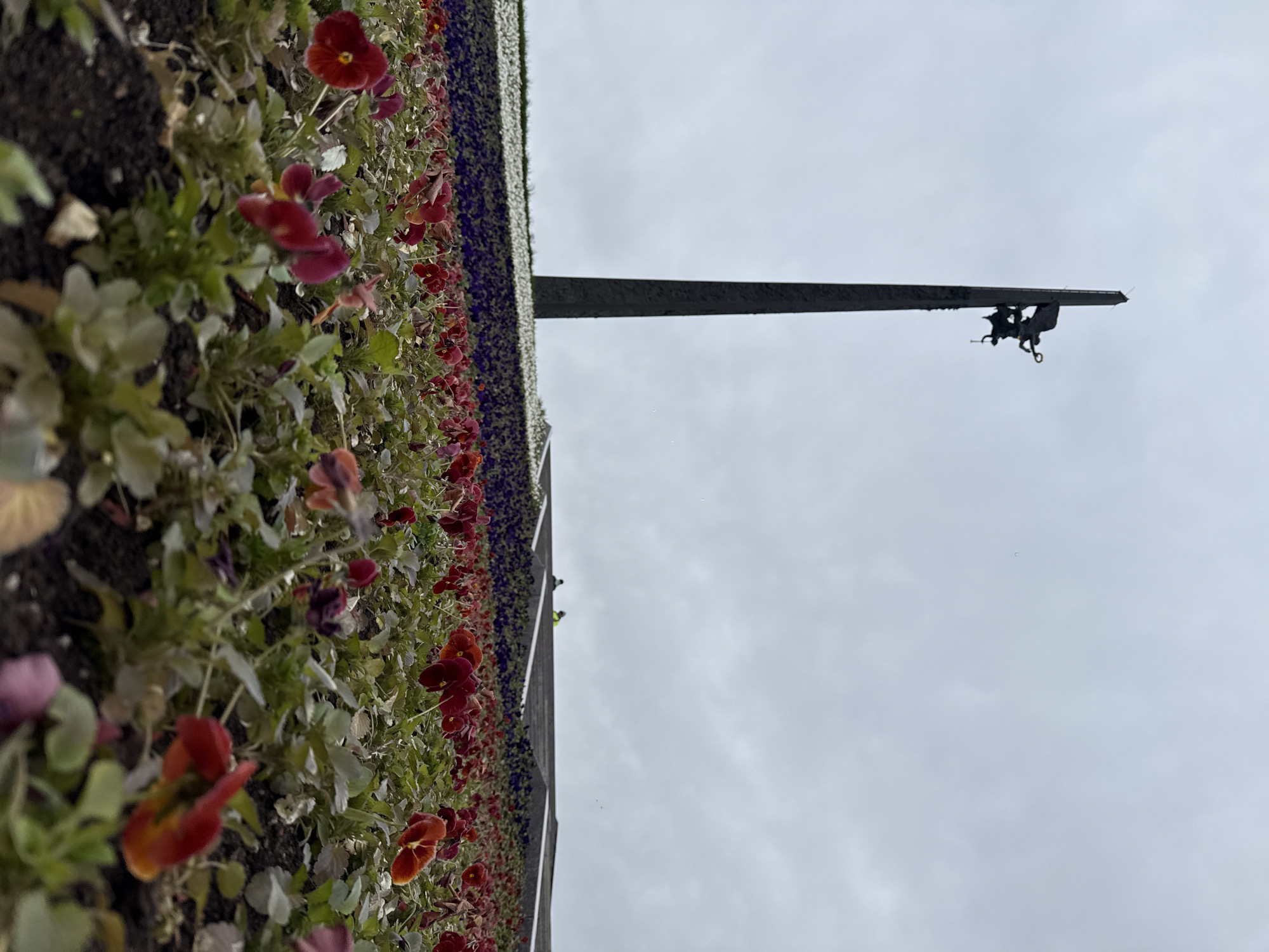 The image captures a scene in Moscow, Russia, showcasing a flower display lining the base of a structure with a metallic arm extending towards the overcast sky. The flowerbed is a vibrant mix of red, purple, and white pansies. A metal arm, which is part of the Conquerors of Space monument, points upward. Two small human figures are visible at the end of the arm. The background consists of a uniform gray, cloud-filled sky.