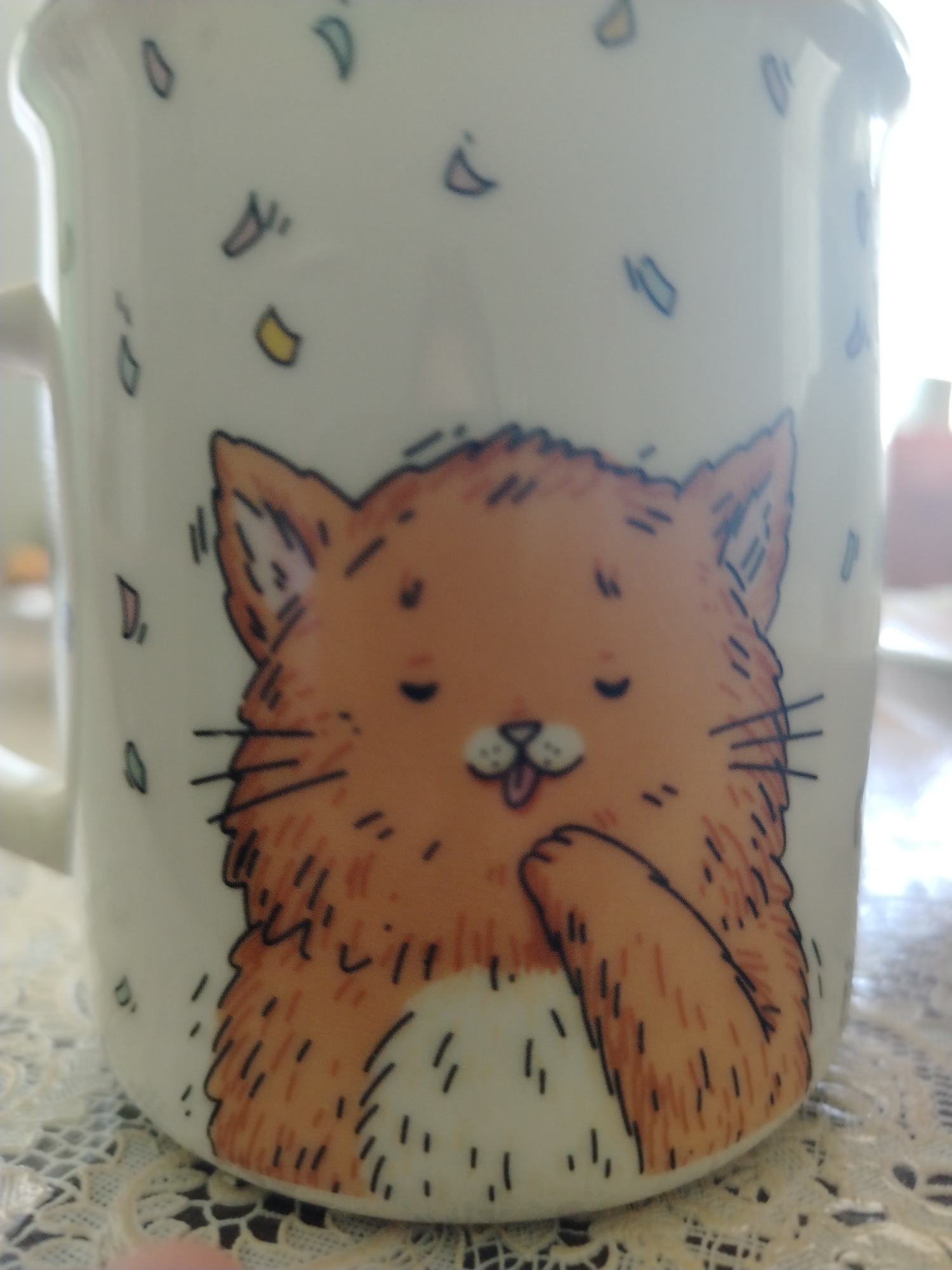 The image shows a white ceramic mug with an illustration of a cartoonish ginger cat. The cat has a fluffy mane, closed eyes with small black dots, a small pink tongue sticking out between its teeth, and white paws. The mug is decorated with colorful confetti-like shapes in pastel colors. The mug is placed on a lace doily. The background is blurred, but appears to be an indoor setting with a window allowing light to enter. The image does not contain any information or context that would suggest its location in the city of Shaki, Azerbaijan.