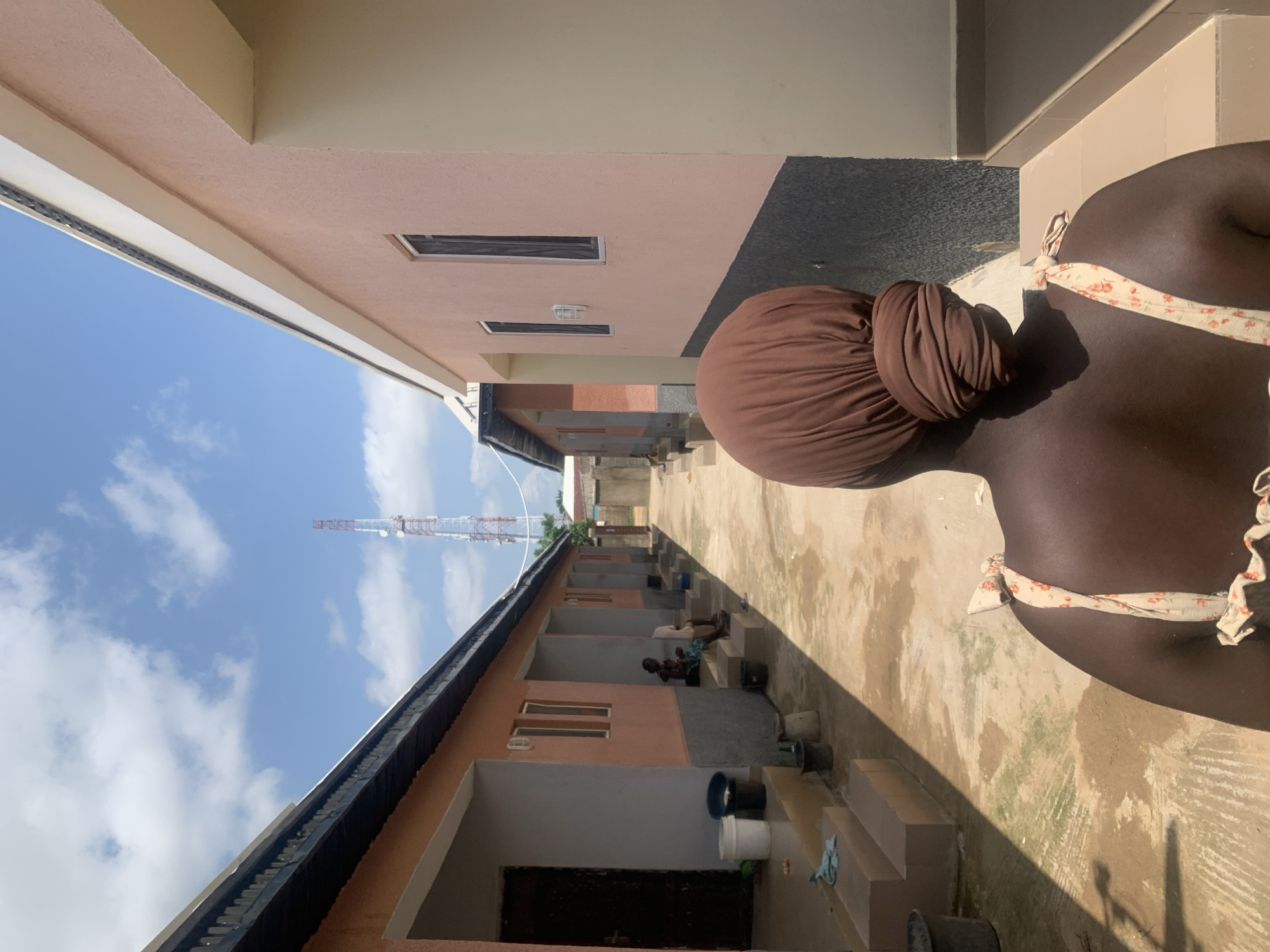 The image depicts an outdoor scene in Jalingo, Nigeria, featuring a narrow walkway between two rows of residential buildings. The buildings are painted in pastel colors, predominantly pink and peach, with dark gray accents near the ground. White trim lines the roofs of the buildings. On the left side of the walkway, there are doorways and windows visible, along with steps leading up to the entrances. Several buckets and containers are placed near the steps. Further down the walkway, two individuals are seated on the steps of one of the buildings.
On the right side of the walkway, the pink-colored building has several windows, some with screens. The walkway itself is paved with concrete that appears worn and has some patches of dirt or discoloration. In the background, a tall telecommunications tower with a lattice structure is visible against a bright blue sky with scattered white clouds. The overall impression is one of a simple, residential area in a city.
The foreground of the image is dominated by the back of a person wearing a brown headwrap and a light-colored, floral-patterned top with a tie strap on the shoulder. This individual is facing away from the camera, looking down the walkway.