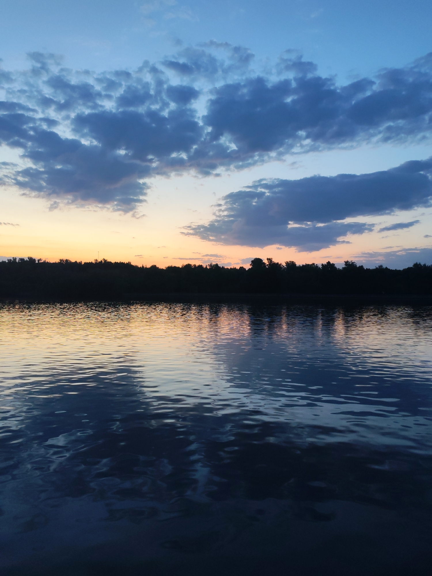 The image captures a twilight scene over a body of water, likely a lake or a wide river. The sky is a gradient of blue at the top, transitioning to orange and yellow near the horizon. Clouds are scattered across the sky, with darker, bluish-gray formations dominating the upper portion.
A dark, silhouetted treeline stretches across the horizon, separating the sky from the water. The water surface is rippled, reflecting the colors of the sky and the trees, creating a slightly blurred and mirrored effect.
There are no visible people or other identifiable objects in the scene. The setting appears to be a natural environment, possibly on the outskirts of Ermolino, Russia, though specific landmarks are not present to confirm this definitively. The focus is on the natural landscape and the interplay of light and reflection.