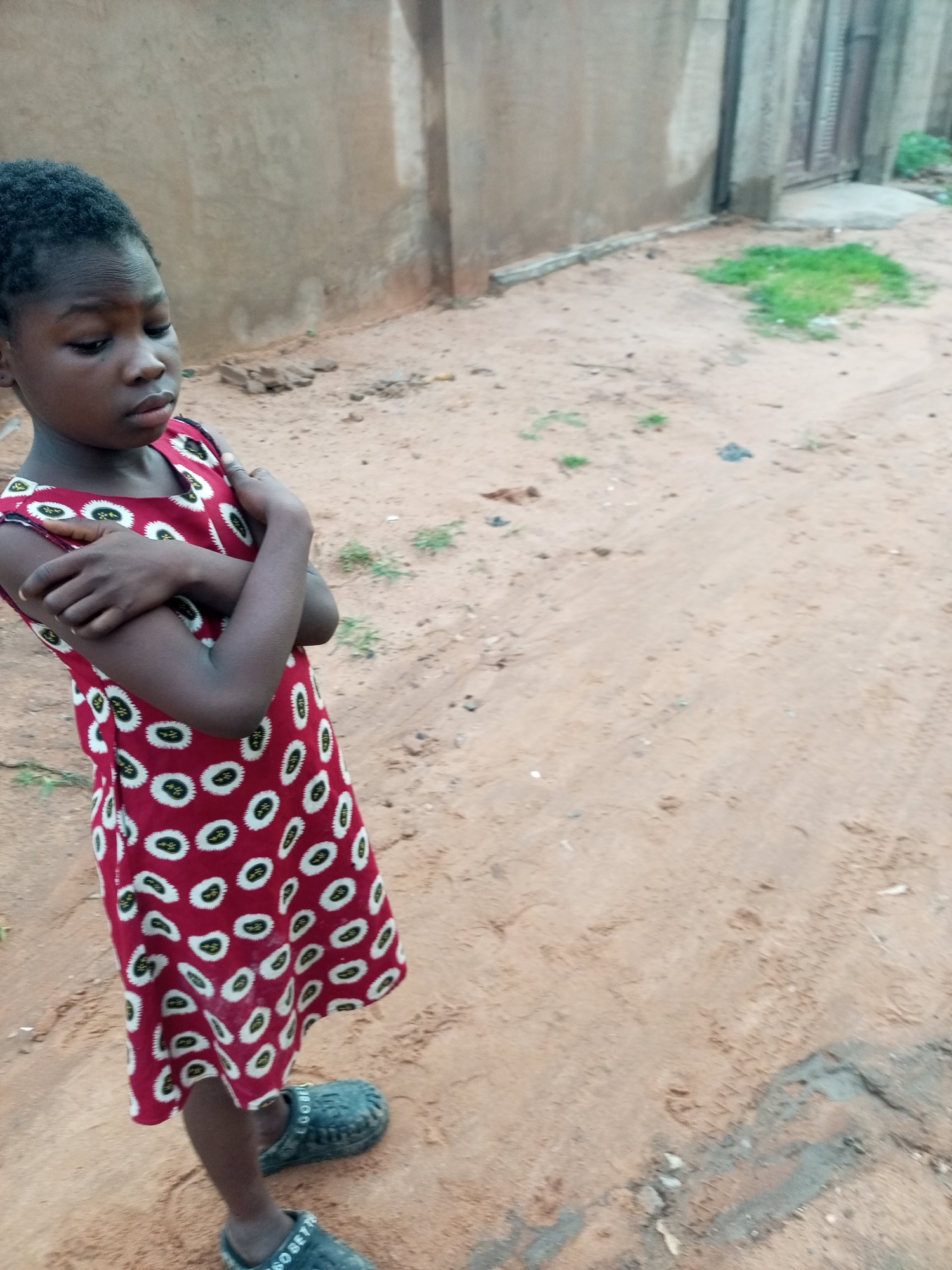 A young Black child stands with their arms crossed, looking down, in a dusty outdoor environment. The child is wearing a red dress with a white and black oval pattern and dark, clunky slip-on shoes. Behind the child is a weathered, light brown wall, and to the right of the child, the ground is sandy with patches of sparse green grass and some debris. The overall impression is of an unpaved area in a residential or less developed part of a city, consistent with a location like Tempure, Nigeria.