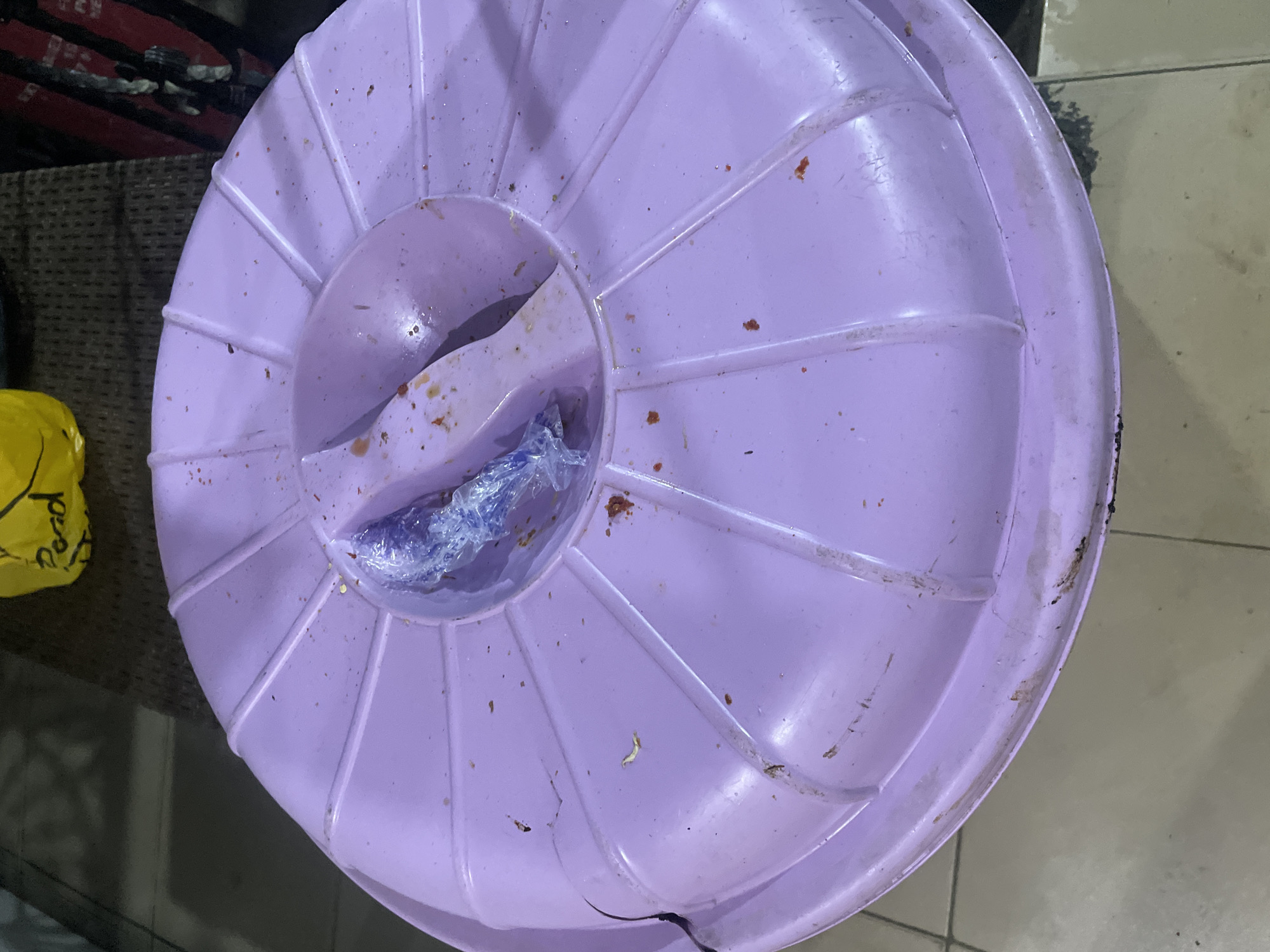 A light purple, round plastic lid with radiating ridges sits on a tiled floor. A raised, oval-shaped opening in the center of the lid contains a crumpled piece of clear plastic wrap with a blue object inside. Small reddish-brown stains and specks are visible on the surface of the lid. In the upper background, a crumpled yellow plastic bag with writing is visible. The environment appears to be indoors.