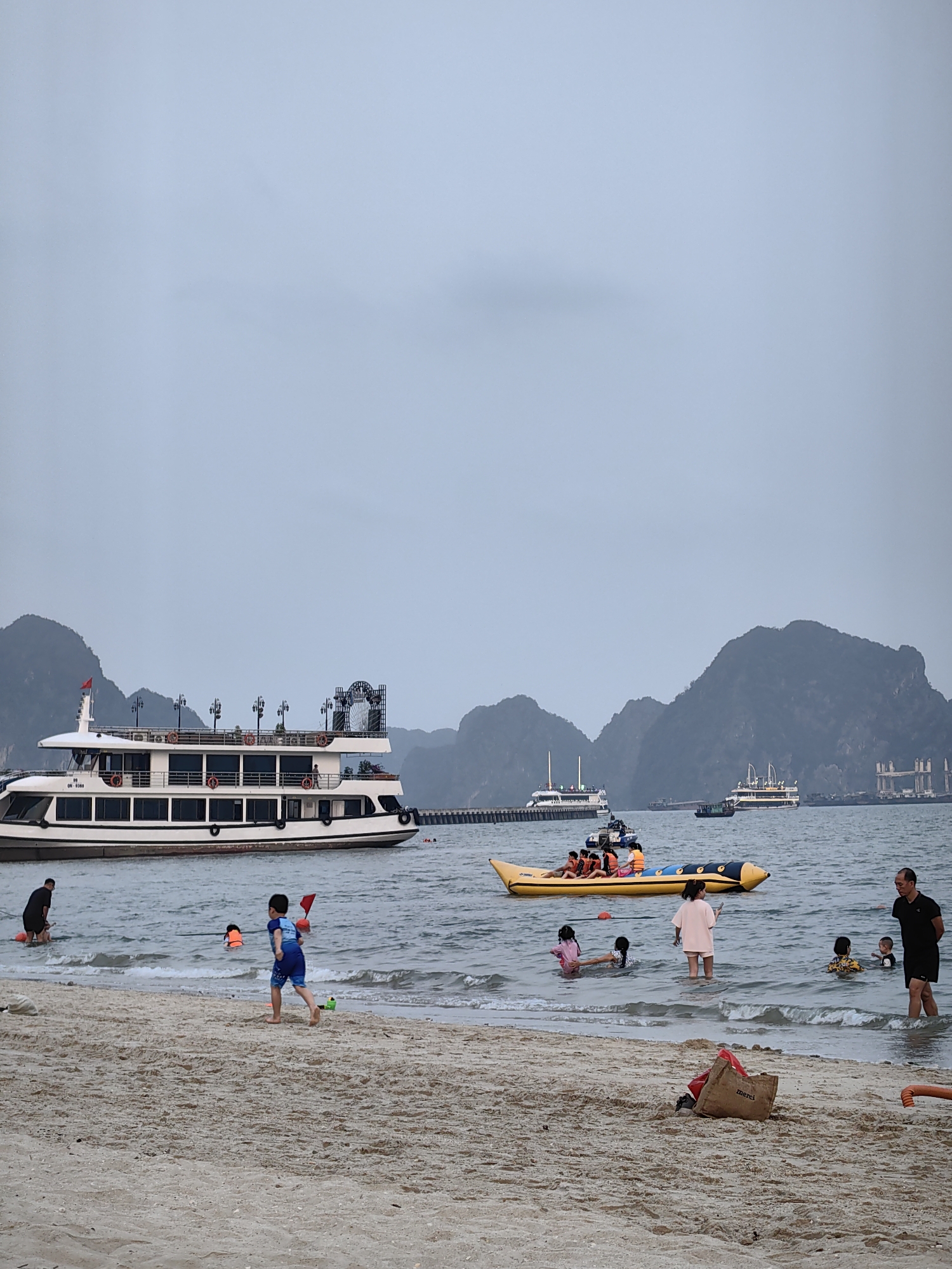 The image shows a beach scene in Bai Chay Ward, Vietnam. The sky is overcast. In the background, there are several islands and a cruise ship docked near a pier. In the water, a banana boat with several people wearing life jackets is visible. Closer to the shore, several people are wading and swimming in the water. On the beach, a young boy in a blue swimsuit is running and holding a red toy. A bag is lying on the sand. An adult stands in the water near the shore. Other people are scattered in the water.