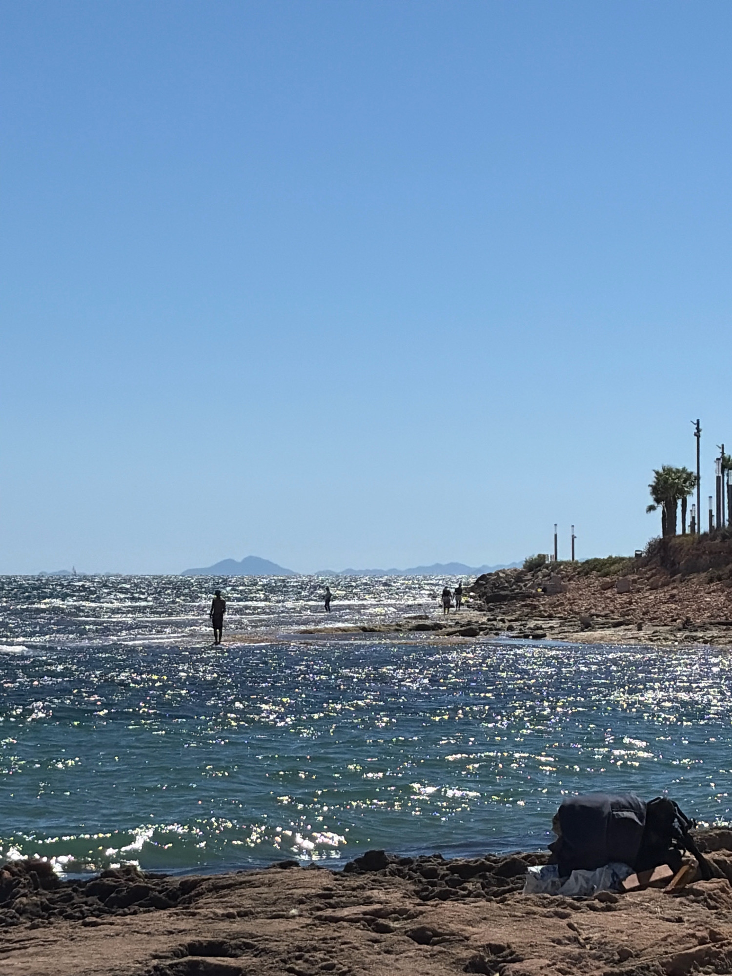 The image captures a sunlit beach scene in Torrevieja, Spain. The foreground is dominated by a rocky shoreline with a dark backpack and clothing laid out on the stones. The mid-ground features the shimmering, sun-dappled surface of the Mediterranean Sea, with several silhouetted figures wading or walking through the shallow water. In the distance, a chain of low, mountainous islands rises against a clear, bright blue sky. Palm trees and lampposts are visible along the coastline on the right side of the image, suggesting a developed beachfront area. The overall impression is one of a peaceful, sunny day at the coast.