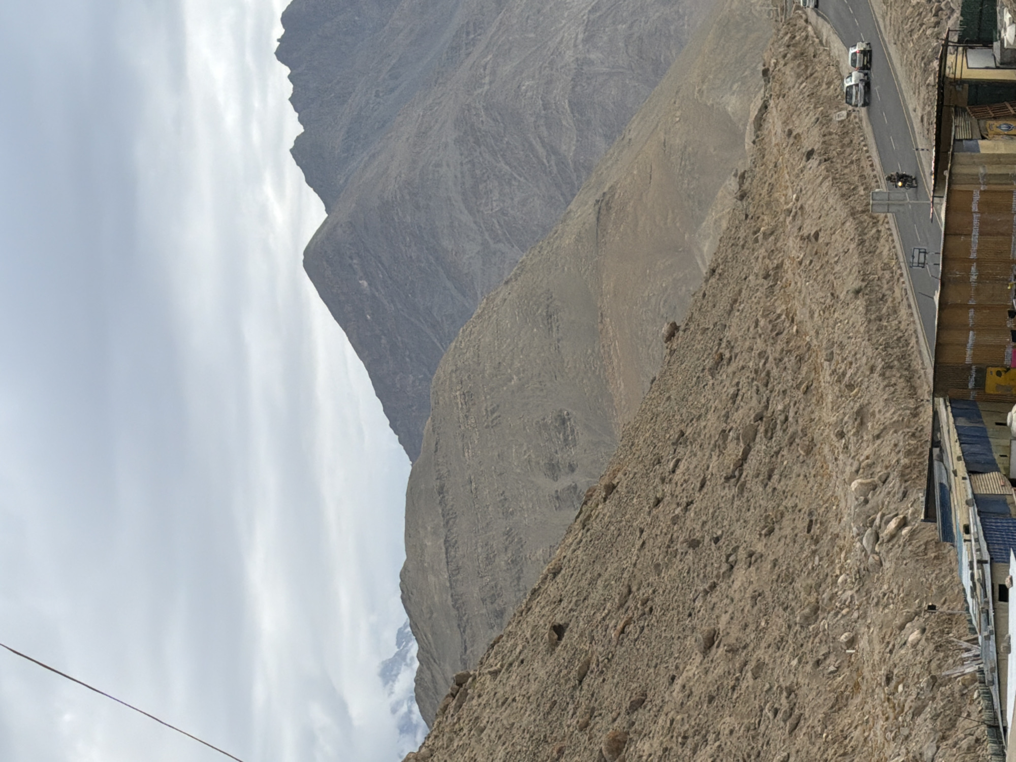In Chumkar, India, a mountainous landscape features a road with vehicles. A string stretches across the cloudy sky from the upper left corner of the image. To the left, a steep, rocky hill rises, covered in loose stones and dirt. Further back, a wider mountain range with layered rock formations dominates the background. A paved road curves through the scene, with two white cars and a motorcycle visible. In the foreground, a low-rise structure with a corrugated metal roof and various walls is partially visible on the right. The sky is overcast with diffused light.