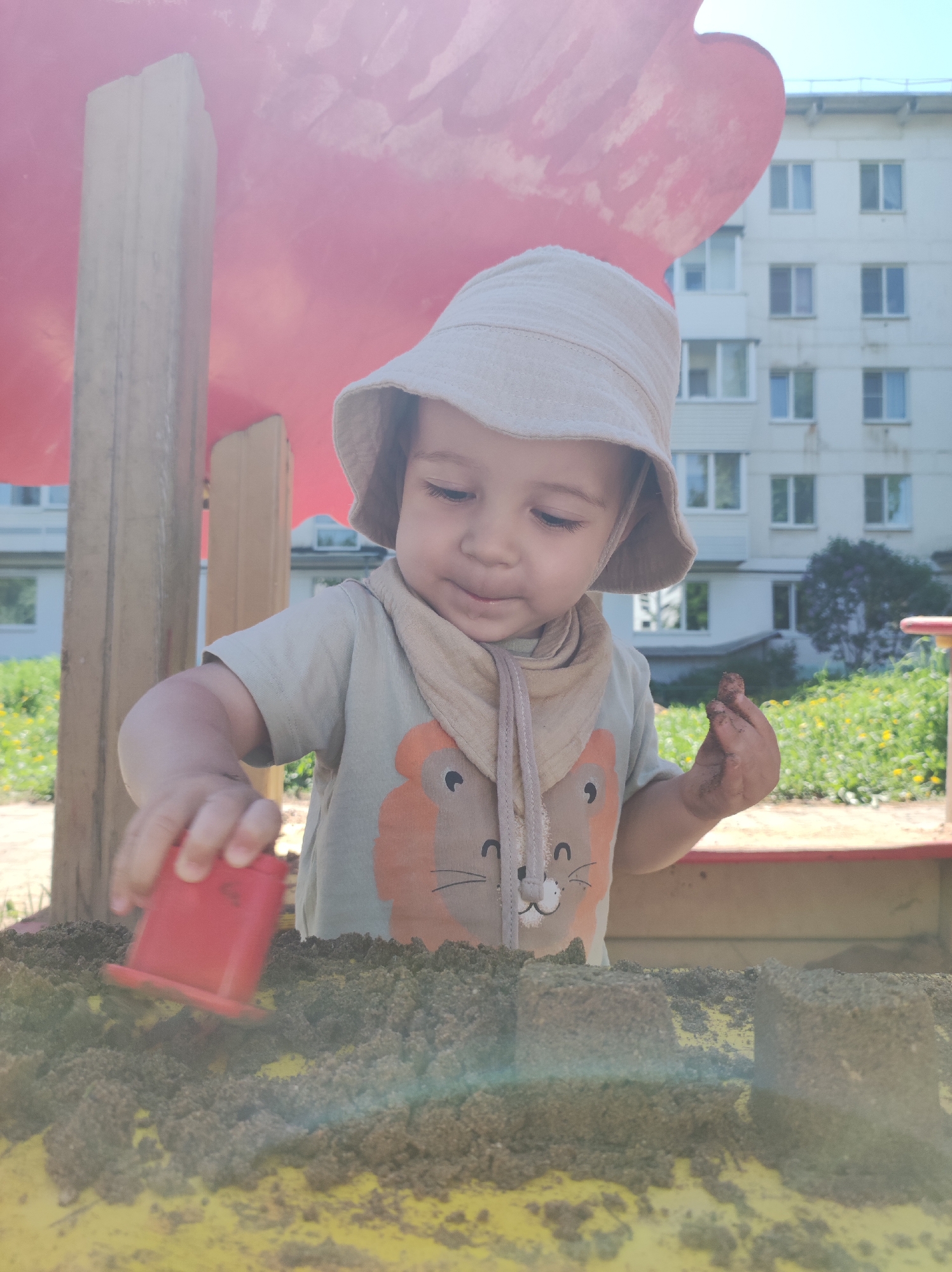 A young child is playing in a sandbox. The child, wearing a light-colored sun hat and a t-shirt with a cartoon lion design, is holding a red plastic cup over a mound of dark sand. The child's right hand is covered in dirt. The sandbox appears to be yellow. Behind the child, there's a red painted structure, likely part of a playground. In the background, a multi-story apartment building is visible, along with patches of green grass and foliage. The photograph was taken in the city of Chaikovsky, Russia.