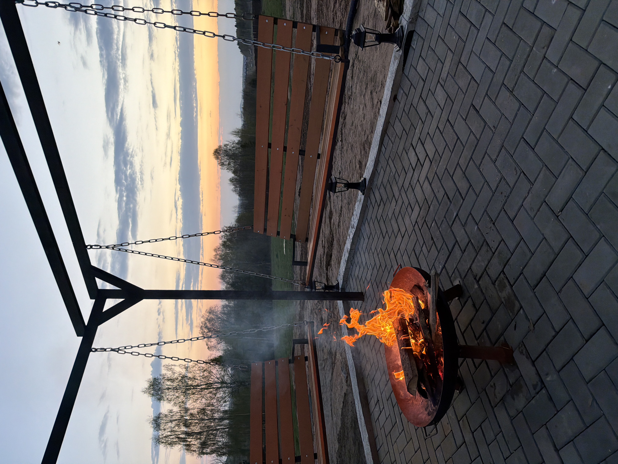 The image depicts a fire pit with flames burning brightly in the foreground, positioned on a paved surface of dark gray bricks laid in a herringbone pattern. To the left, a swing set structure with two wooden benches hanging from chains is visible. The backdrop features a landscape with trees and a sky illuminated by a setting sun, exhibiting a gradient of colors from pale blue to orange and pink hues. The image gives the location in the city of Kamennoye, Russia. There are no people visible in the scene.