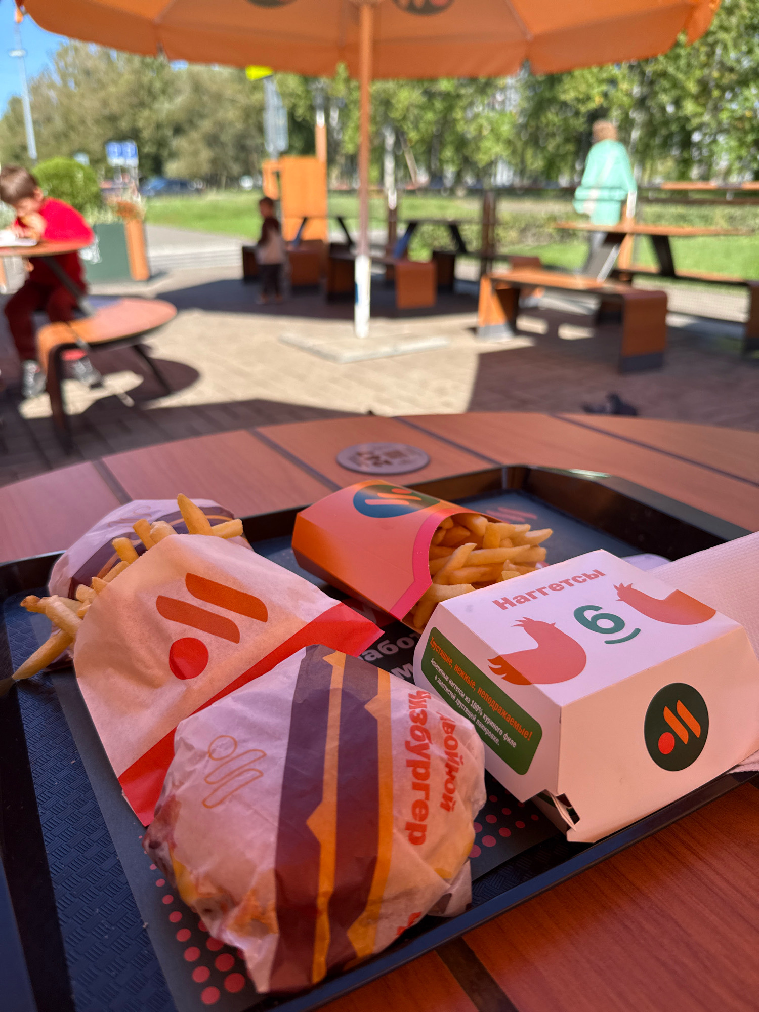 The image shows a meal of french fries and chicken nuggets from McDonald's, placed on a tray on an outdoor table. In the background, several children are visible in a park-like setting. One child in a red shirt is seated at a table, while another child in a light blue shirt stands near a picnic table. The overall scene suggests a casual dining experience outdoors on a sunny day. The text on the chicken nugget box is in Russian, reading "Наггетсы 6" (Nuggets 6). The location is identified as Rusanovo, Russia.