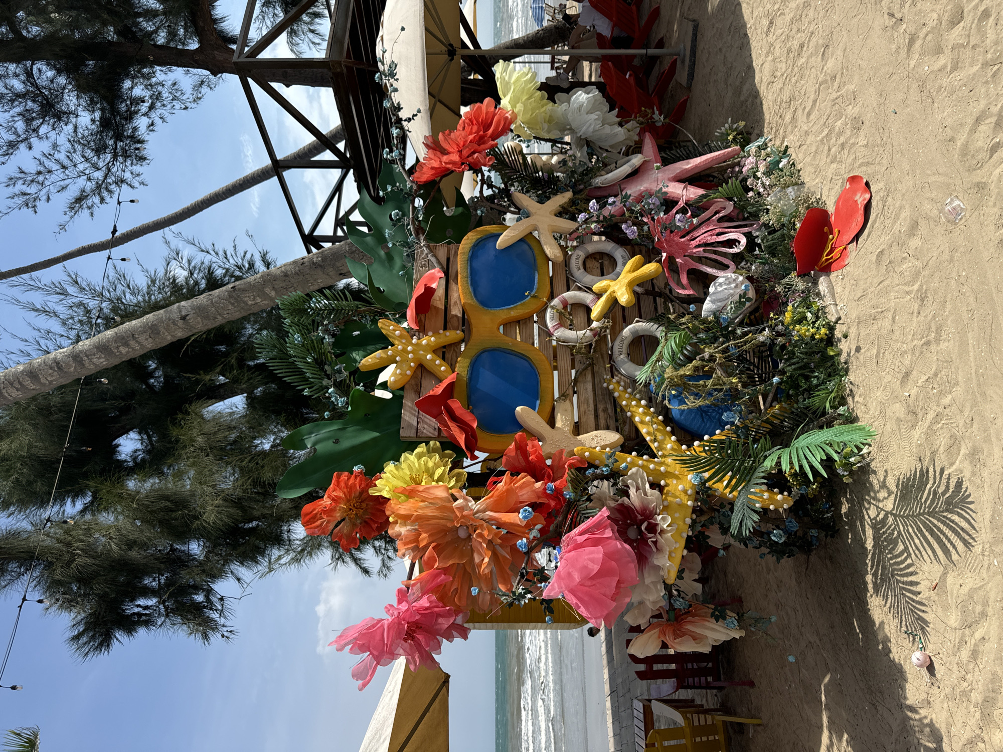 This media file depicts a vibrant, beach-themed decoration, likely at a resort or cafe in Mũi Né, Vietnam. The primary subject is a colorful, artificial floral arrangement adorned with nautical and tropical elements. Prominently featured are oversized, decorative sunglasses with bright blue lenses, a yellow starfish, and a pink octopus. Several life preserver rings, some with "Welcome" printed on them, are also incorporated into the display. Various faux flowers in shades of red, orange, yellow, pink, and white, along with green palm fronds and smaller blue floral accents, create a lively, summery aesthetic.
The scene is set against a backdrop of sandy beach and the ocean, with a clear blue sky and some clouds suggesting it is daytime, likely sunny given the strong shadows. In the background, a tall palm tree trunk and branches, along with parts of a wooden structure and a beach umbrella, hint at a coastal establishment. The overall impression is one of a cheerful and inviting holiday atmosphere, characteristic of a beach destination. There is no visible activity or people in the frame.