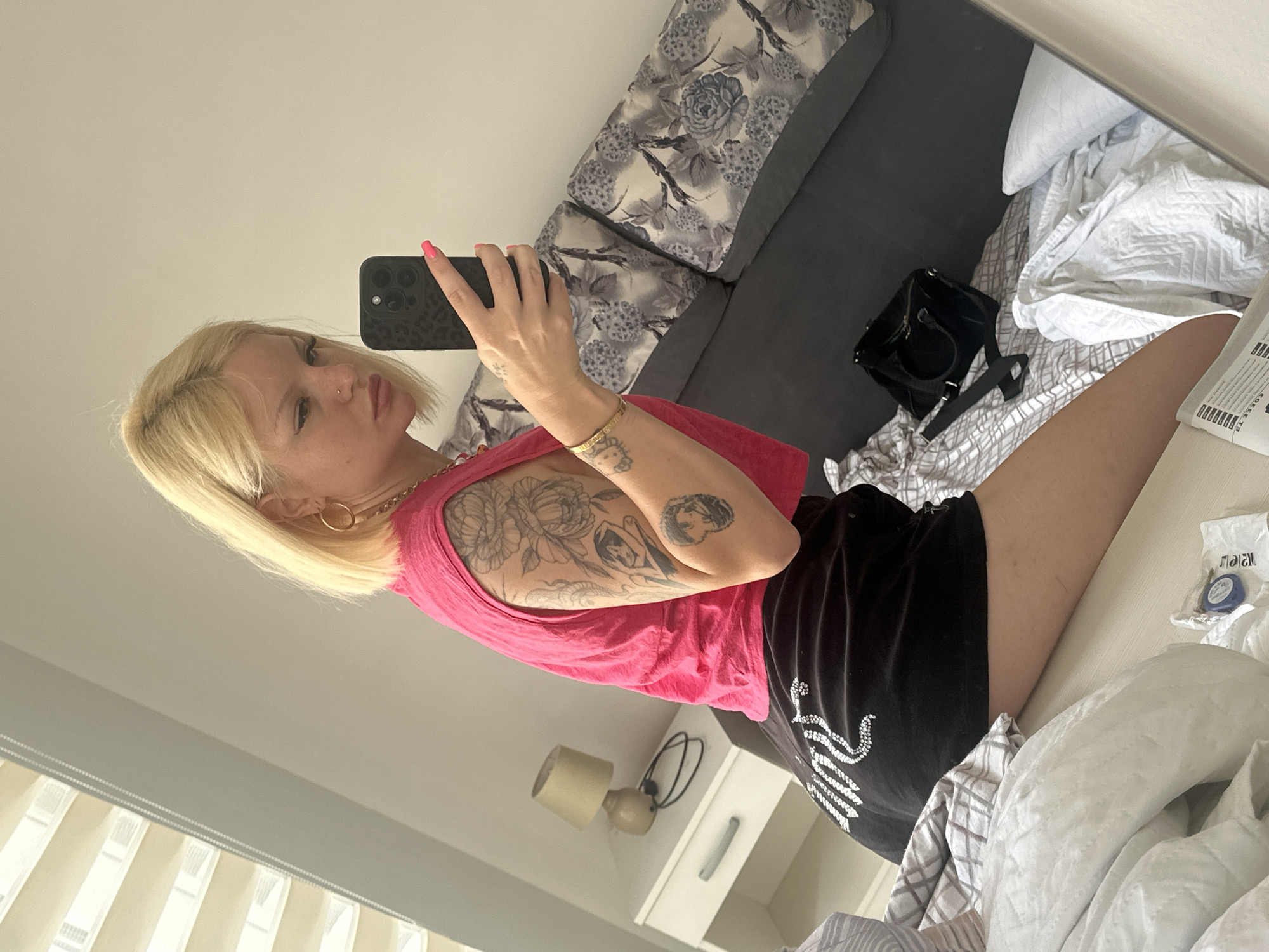 A young blonde woman with tattoos on her arm is taking a mirror selfie in a bedroom. She is wearing a pink sleeveless top and black shorts. The room appears to be in Budva, Montenegro, as indicated by a previous prompt. The bed is unmade, and there are several pillows and blankets scattered around. A nightstand with a lamp is visible to the left of the frame. A black handbag is on the bed near the woman. The blinds on the window are partially open, allowing light to filter into the room.
