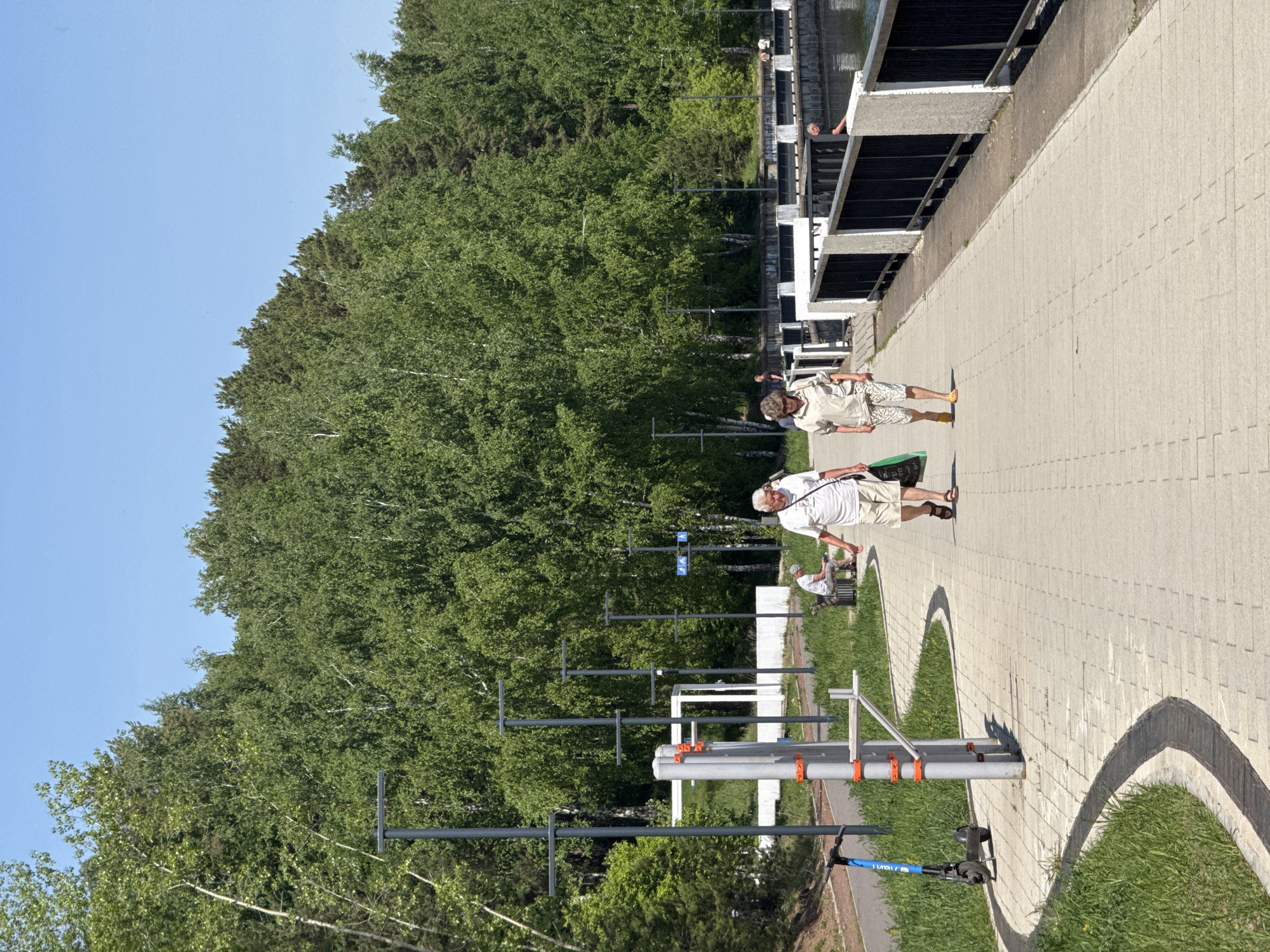 The photograph depicts a partially shaded outdoor scene on a paved walkway, presumably in a park setting within the city of Unknown, Unknown. The walkway, constructed with light-colored rectangular pavers, runs alongside a thick forest on the left. Several metal structures, possibly lamp posts or signage supports, are positioned between the walkway and the forest.
Two adult males are walking along the path, their posture suggesting a leisurely stroll. The man closest to the camera is wearing a white shirt and tan shorts, while the other man is wearing a tan shirt and shorts. An adult is sitting on a bench on the right of the walkway in front of a grassy area.
A section of the walkway is bordered by a low, dark grey wall or structure, possibly a railing or barrier. The sky above the trees is clear and blue, indicating a sunny day. The overall environment suggests a public recreational area in an urban or suburban setting.