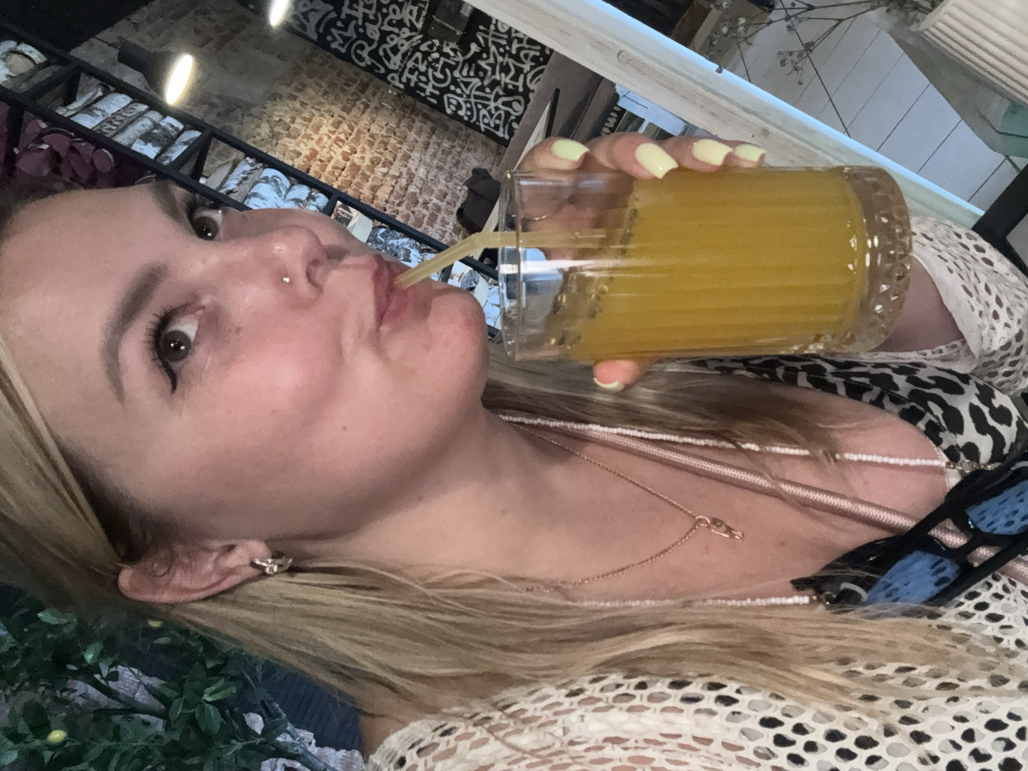 A Caucasian woman with blonde hair and a nose ring is drinking from a tall glass of orange juice with a yellow straw. She is wearing a white crocheted overlay, a leopard print top, and a rose gold pendant necklace. Her fingernails are painted light yellow. The background suggests an indoor setting, possibly a cafe or restaurant, with exposed brick walls and decorative shelving. A plant with small yellow fruits is visible in the upper left corner. The scene is a close-up, focusing on the woman and her drink. No specific location in Chaikovsky, Russia, can be determined from the visual information provided.