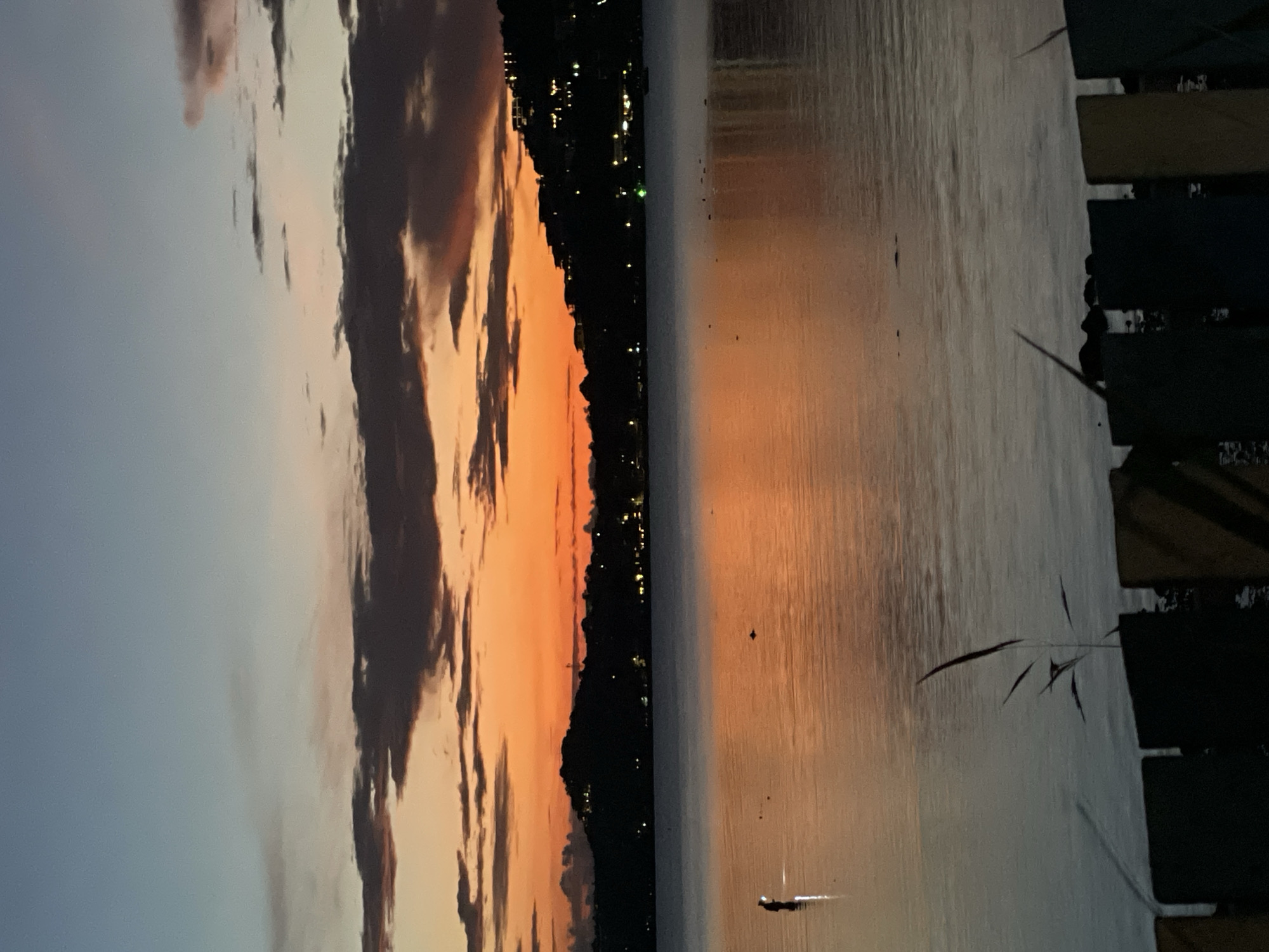 The image depicts a serene twilight scene at Baan Bang Rak, Thailand. A tranquil body of water reflects the vibrant orange and gradient blue hues of the sky, punctuated by dark, silhouetted clouds. Along the distant horizon, a dark, hilly coastline is dotted with illuminated lights, suggesting a settlement. In the foreground, a small section of a wooden fence, partially obscured by a few reeds, borders the water. A solitary figure stands partially submerged in the water, facing the horizon, their silhouette dimly illuminated. The overall impression is one of peaceful dusk over a coastal landscape.