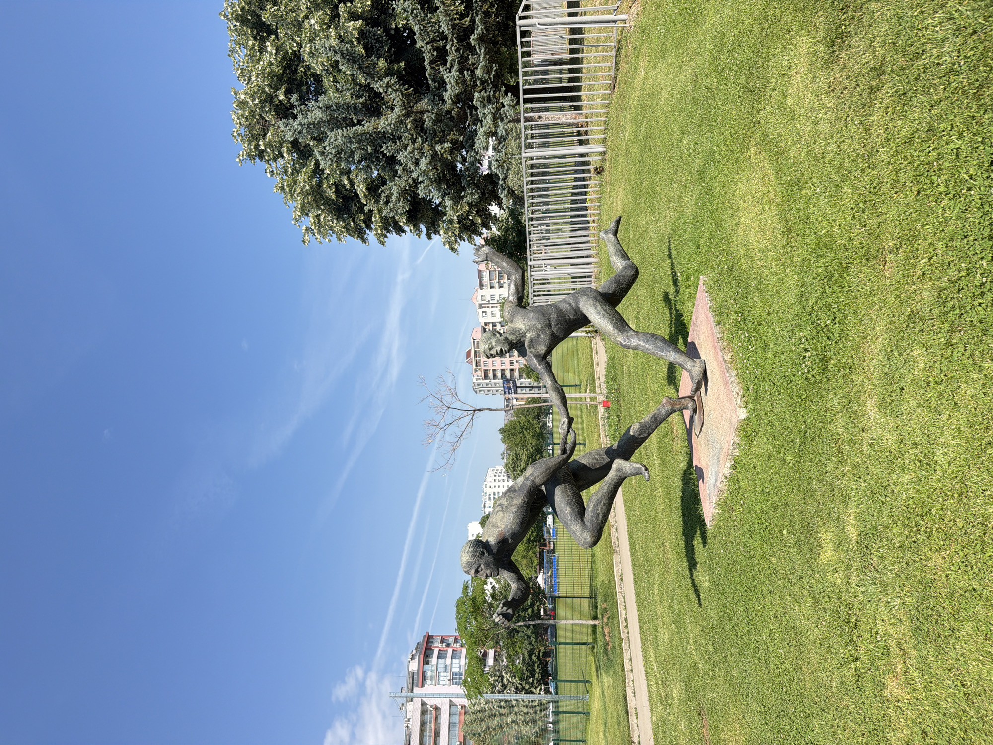 The image depicts a bronze sculpture of two athletes in mid-stride, appearing to be in a race. They are positioned on a concrete base surrounded by a lush green lawn. In the background, several multi-story apartment buildings are visible, characteristic of urban residential areas. A metal fence runs along the edge of the park area. The sky above is clear blue with a few wispy clouds. This sculpture is located in the South Park (Yuzhen Park) in the city of Sofia, Bulgaria, specifically in an open grassy area near the intersection of "Nikola Vaptsarov" Boulevard and "Arsenalski" Boulevard. The sculpture is called "The Runners."