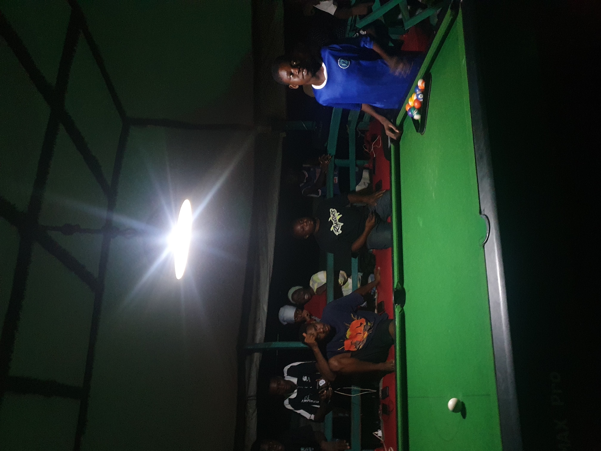 The image depicts a group of young people gathered around a pool table in what appears to be an outdoor or semi-outdoor recreational area. The scene is dimly lit, with a bright overhead light illuminating the pool table and the immediate surroundings. Several individuals are visible in the background, some seated and others standing, observing or participating. In the foreground, a pool table with green felt is set up, with a white cue ball and a rack of colored balls positioned at one end. A young person wearing a blue shirt is standing near the table, seemingly engaged with the game. The overall impression is a casual gathering for a game of pool. The location is identified as Lokoja, Nigeria.